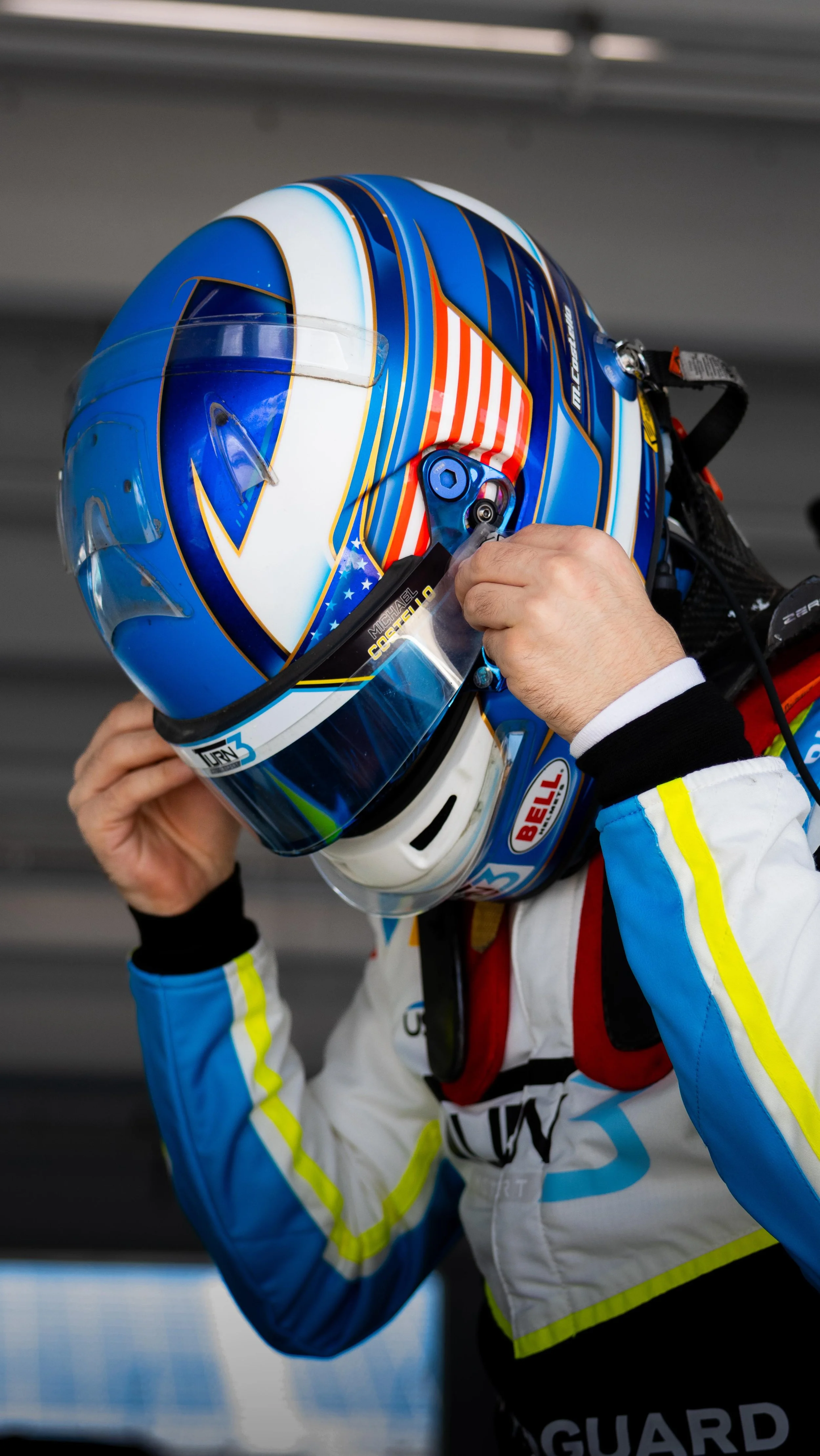 A race car driver wearing a colorful blue, white, and orange helmet, adjusting the helmet strap.