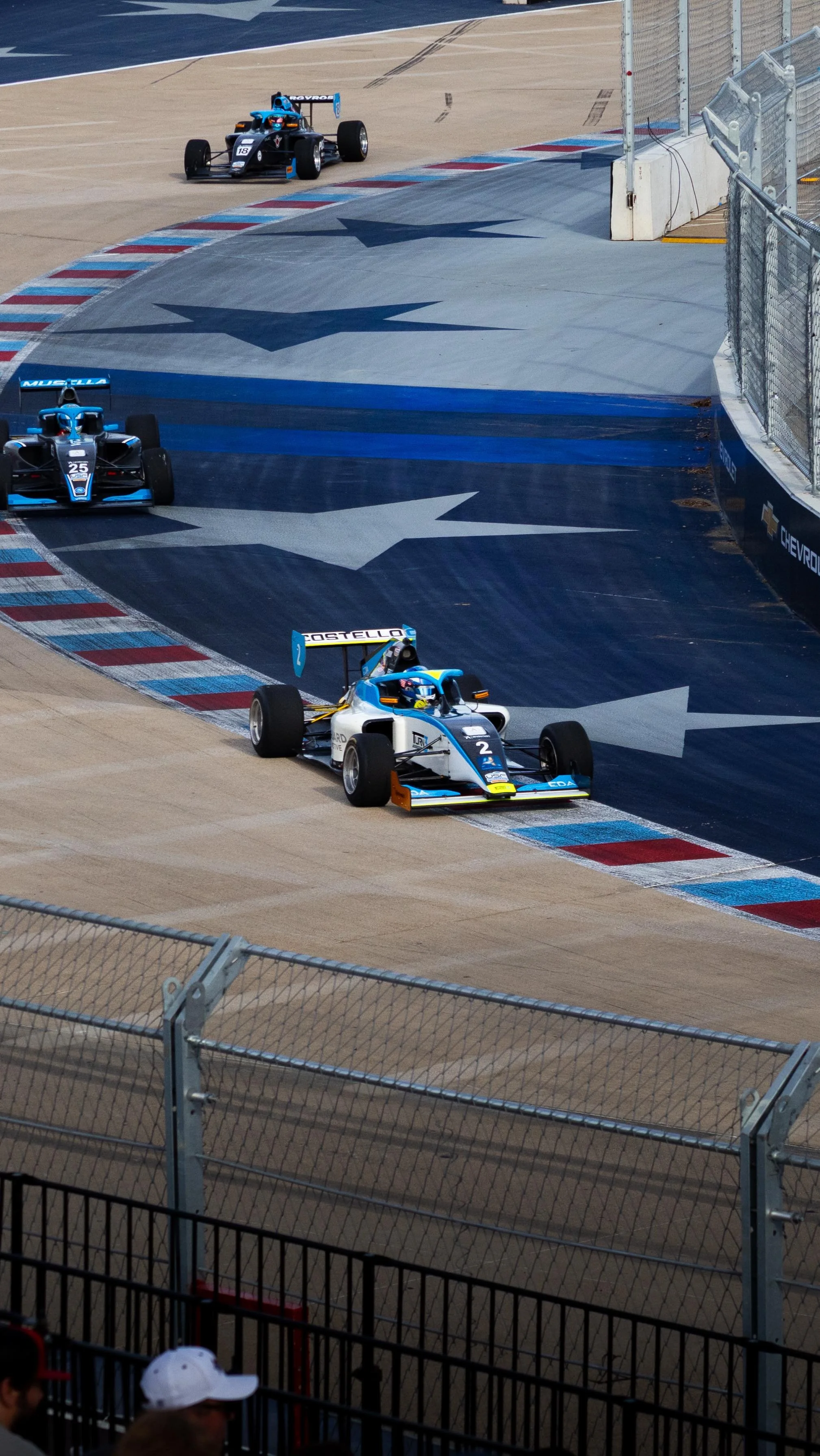 Open-wheel race car on a racetrack near the wall with large painted arrows, with another race car in the distance on the track. In Arlington Texas.