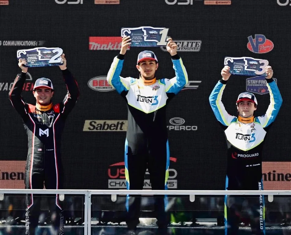 Podium winners holding up their trophies after a race, standing on a winners' podium with a black backdrop covered in logos.