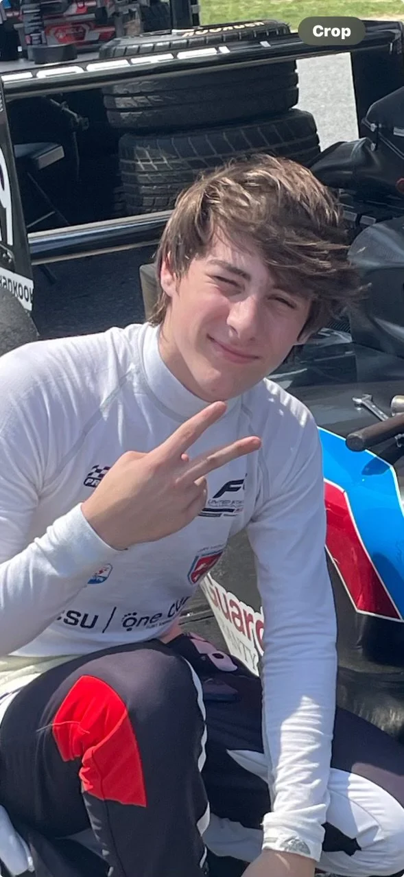 A young man with brown hair making a peace sign, sitting in front of a race car, with a vehicle carrying tires in the background.