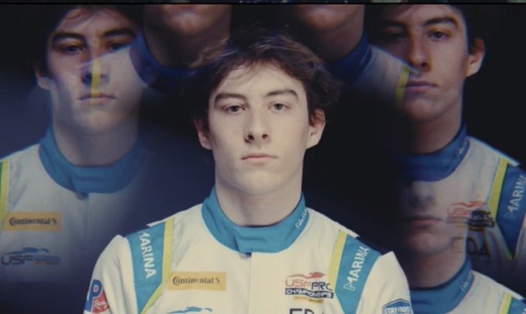 A portrait of a young male race car driver in a racing suit, with multiple faded reflections of himself around him against a dark background.
