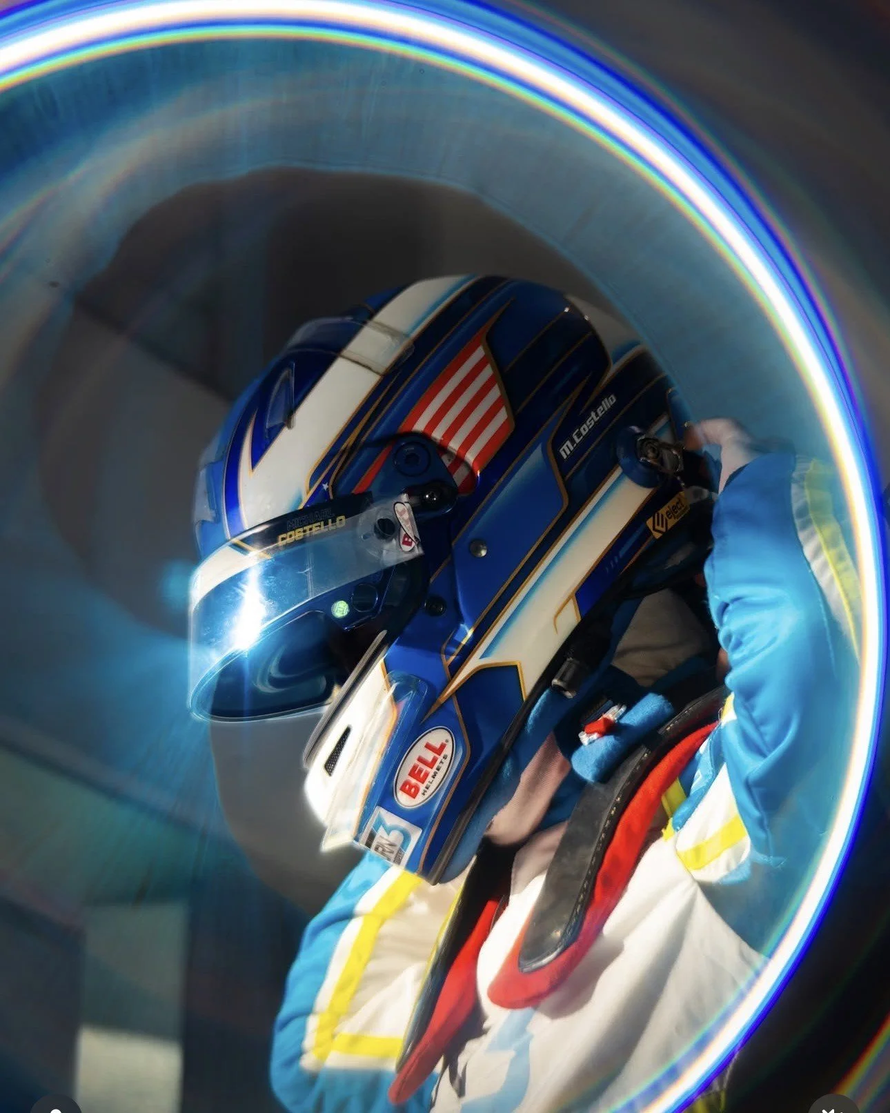 Close-up of a race car driver in a blue, white, and yellow racing suit and helmet inside a calibration tunnel.