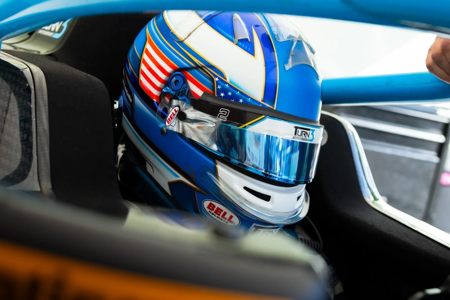 Blue racing helmet with American flag design and racing brand stickers, placed inside a race car.