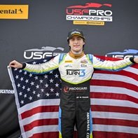Race car driver holding American flags at a podium, wearing a racing suit and a cap, with a sponsorship banner behind him. #Indycar #IndyNXT # USF Pro championship #Champion