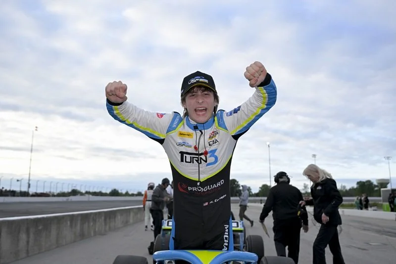 A young male race car driver celebrates with arms raised after a race, wearing a racing suit and black cap, on a race track with several people in the background. #Indycar #IndyNXT # USF Pro championship #Champion