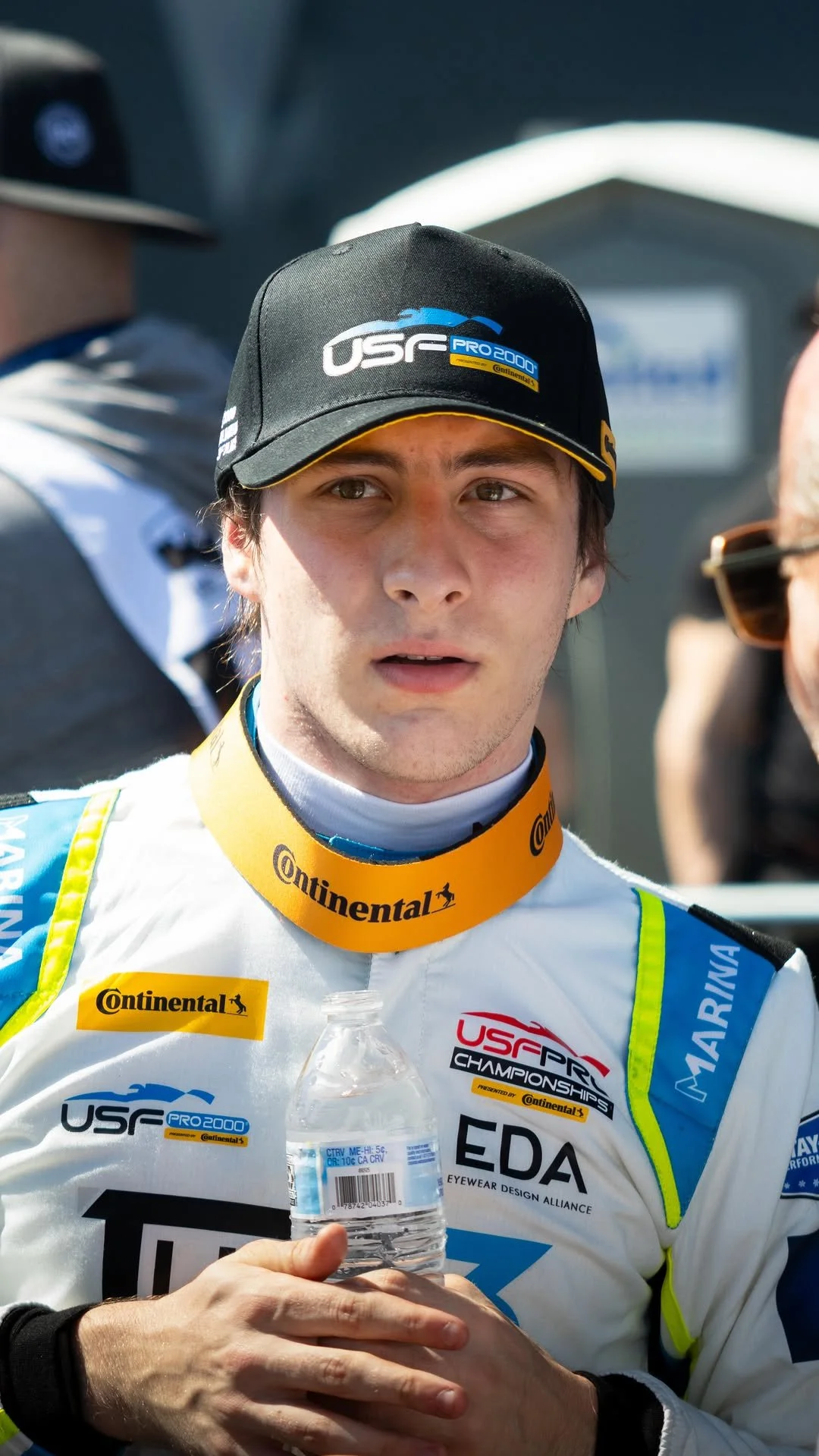 A race car driver holding a water bottle, wearing a white racing suit with various sponsor logos and a black cap, standing outdoors in a sunny environment.