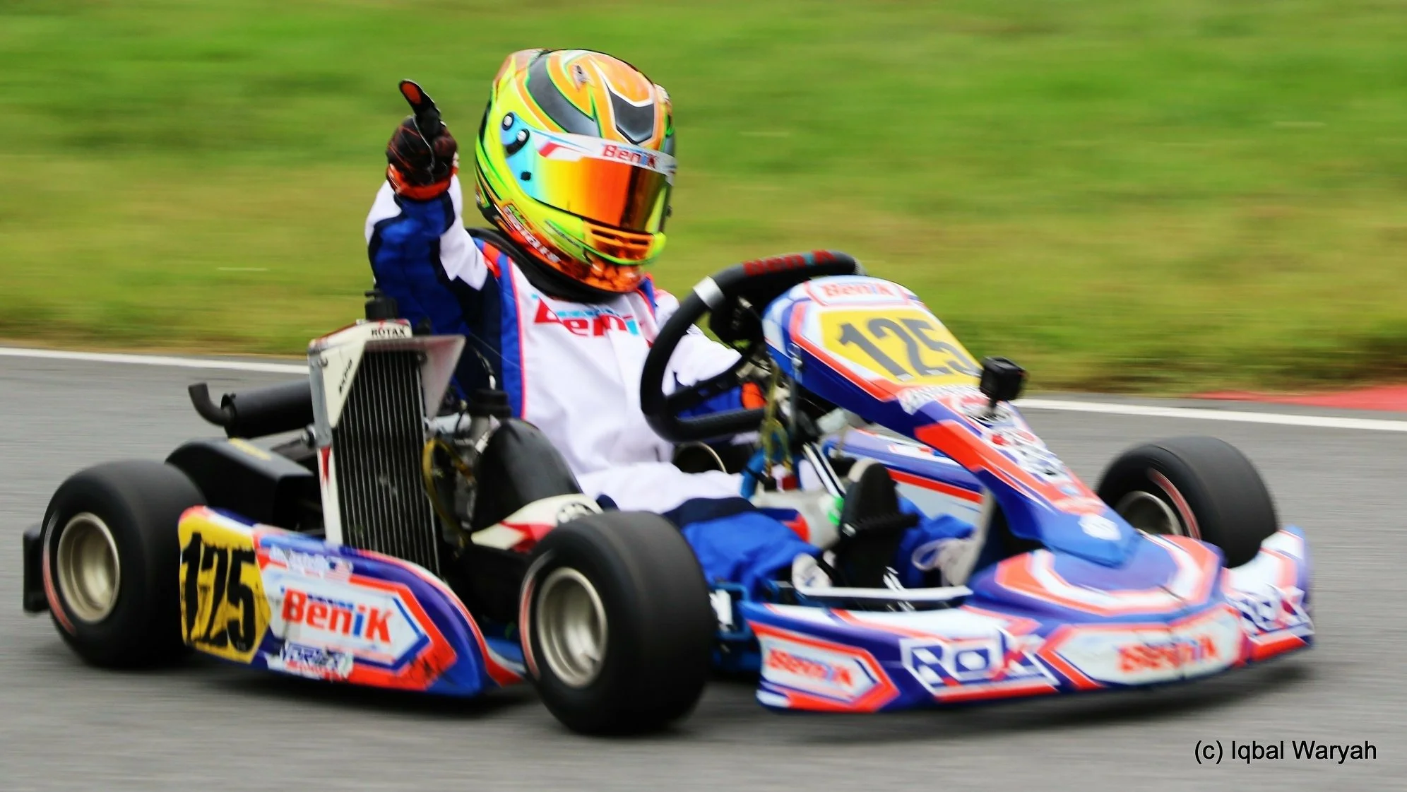 A go-kart racer wearing a colorful helmet and racing suit navigating a turn on the track, with a blurred green background.