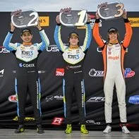 Three race car drivers on the winners' podium holding trophies, with the first and second place drivers on either side, and the third place driver in the middle, all in racing suits. #Indycar #IndyNXT # USF Pro championship #Champion