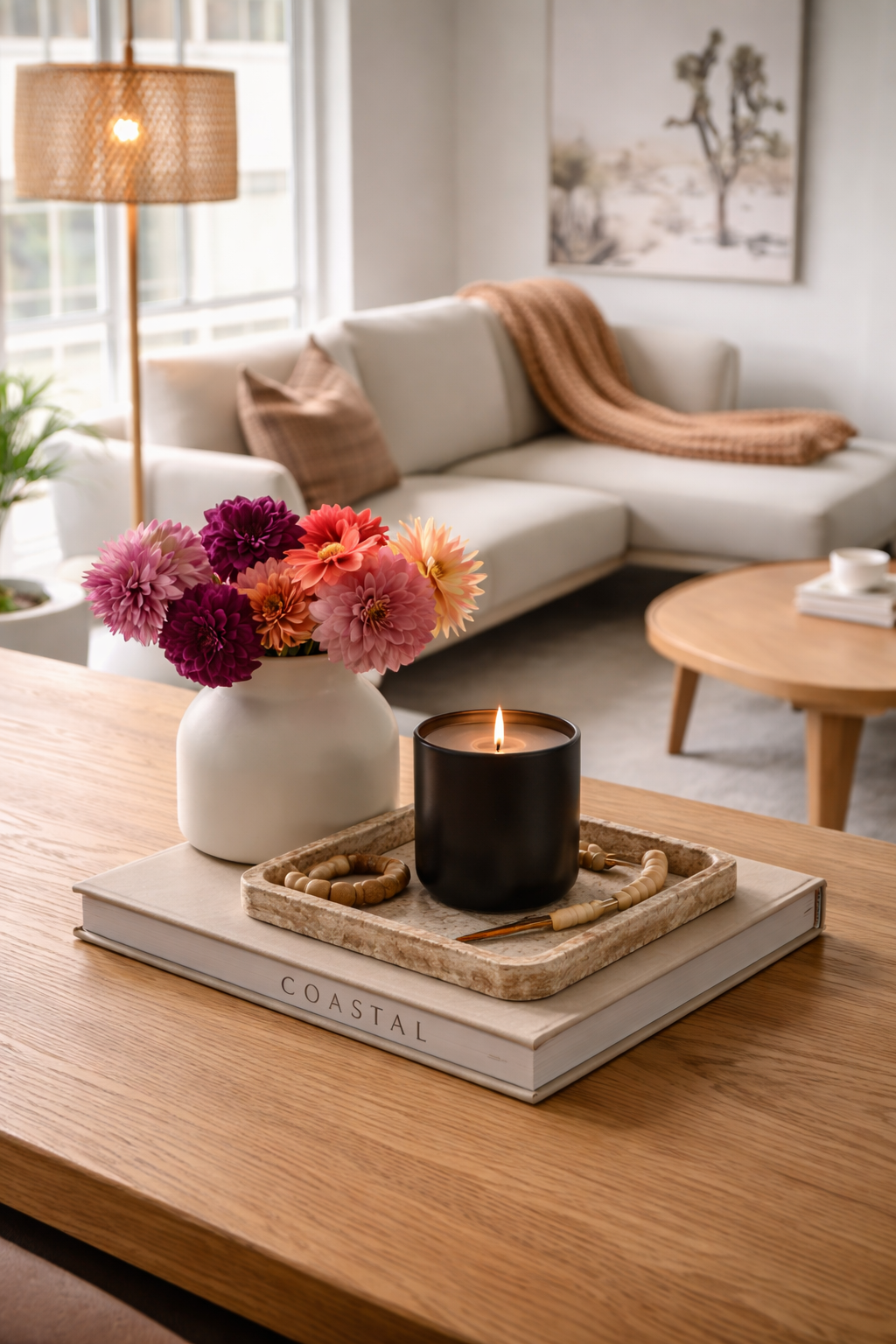 A living room with a white sofa, brown throw pillows, a brown blanket, a painting of a tree, a wooden coffee table with a cup and saucer, a side table, a floor lamp, and a vase of pink, purple, orange, and peach flowers, with a lit black candle on a tray, on top of a large book.