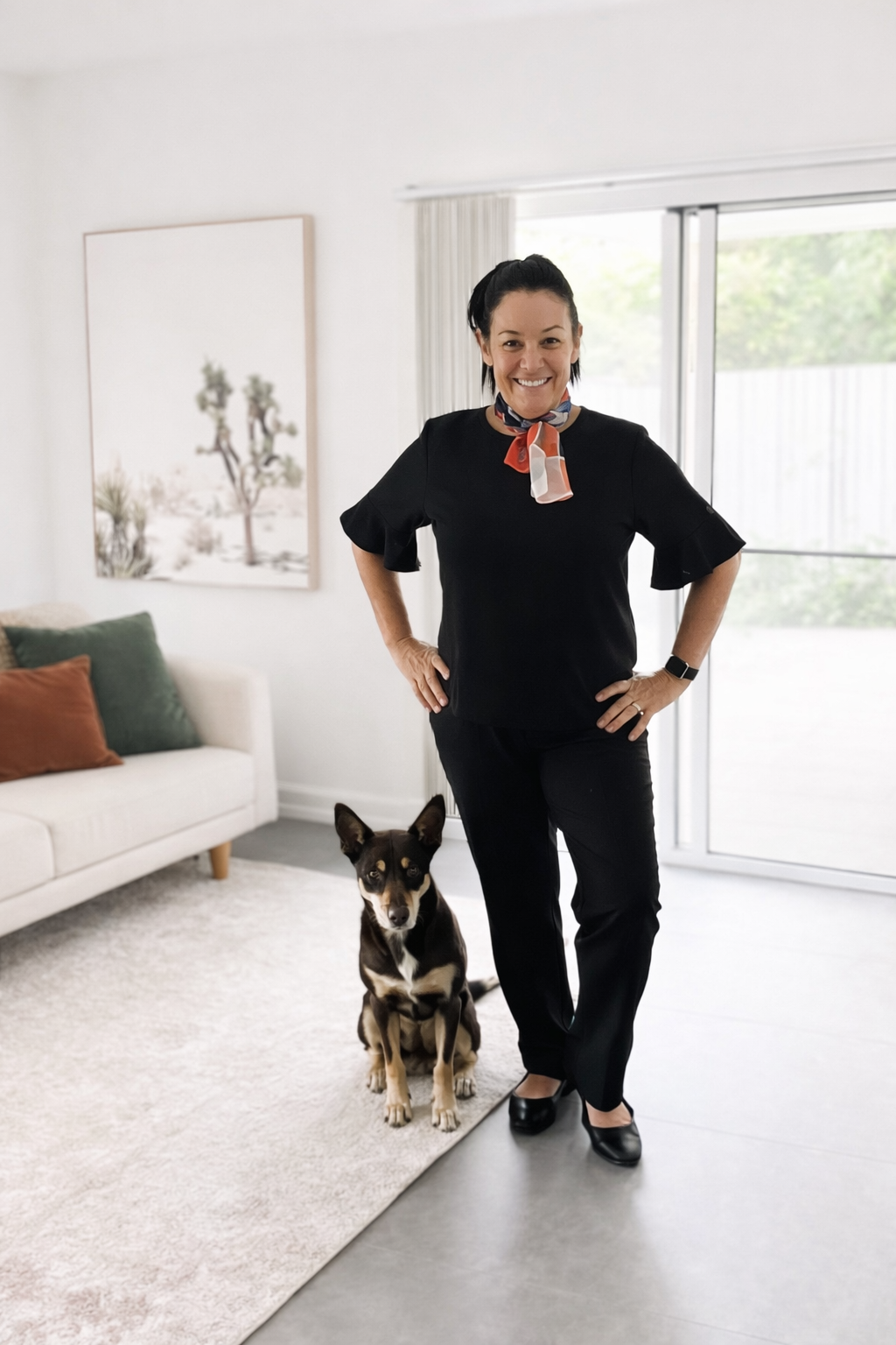 A woman with dark hair in a bun, wearing a black outfit and a red and white scarf, stands in a living room next to a black and tan dog. She is smiling with hands on her hips, and the dog sits attentively beside her. The living room has a white couch with green and orange pillows, a large framed desert landscape artwork, and sliding glass doors with curtains, leading to an outdoor area.