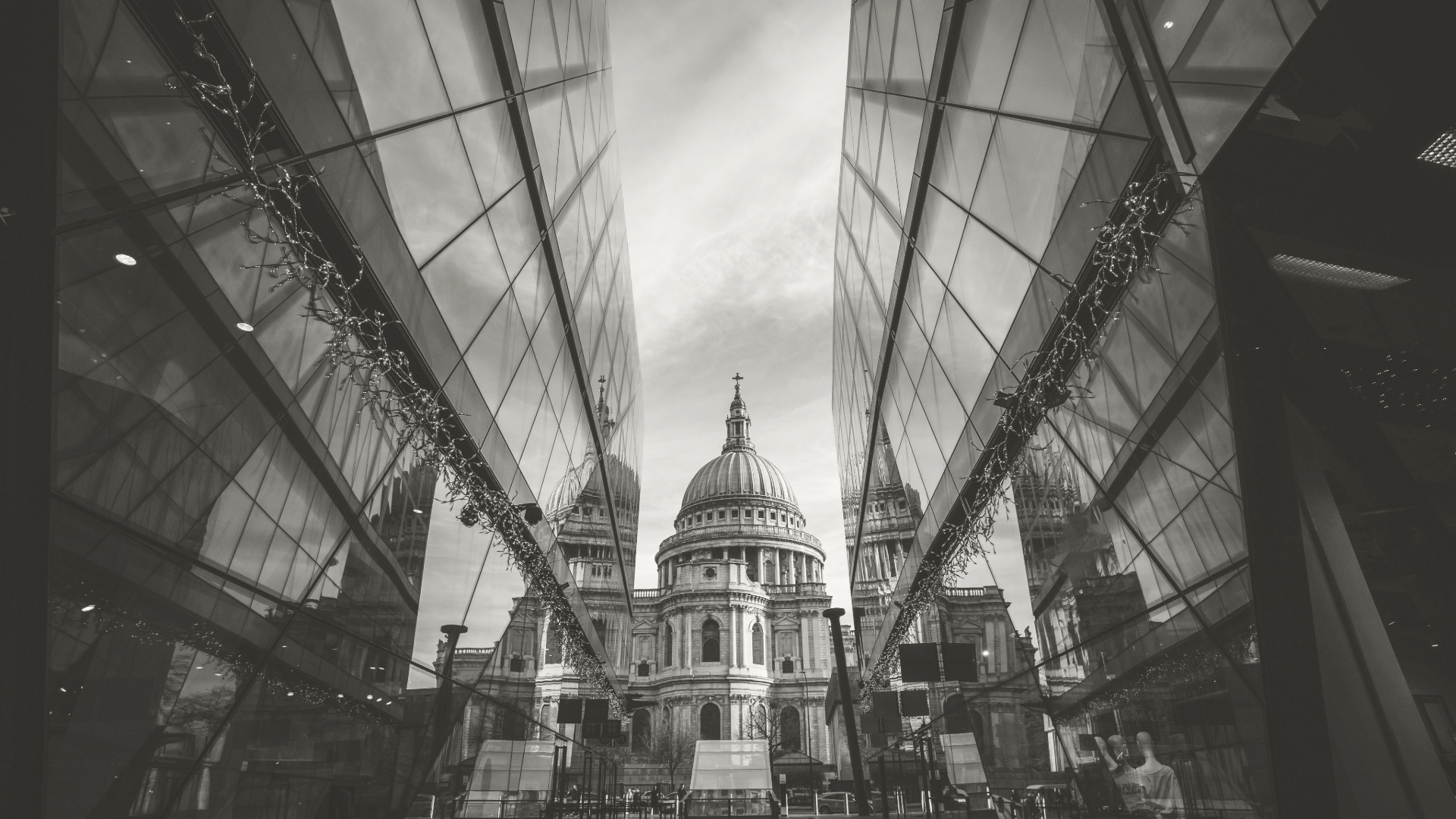 Black and white photograph of St. Paul's Cathedral in London, viewed through a symmetrical glass building with reflections and string lights overhead.