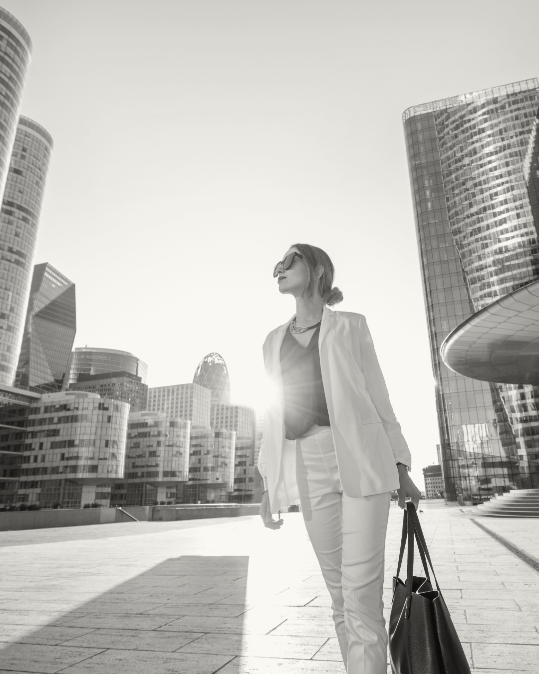 A stylish woman in a suit and sunglasses walking in a modern city with tall buildings in the background during the daytime.