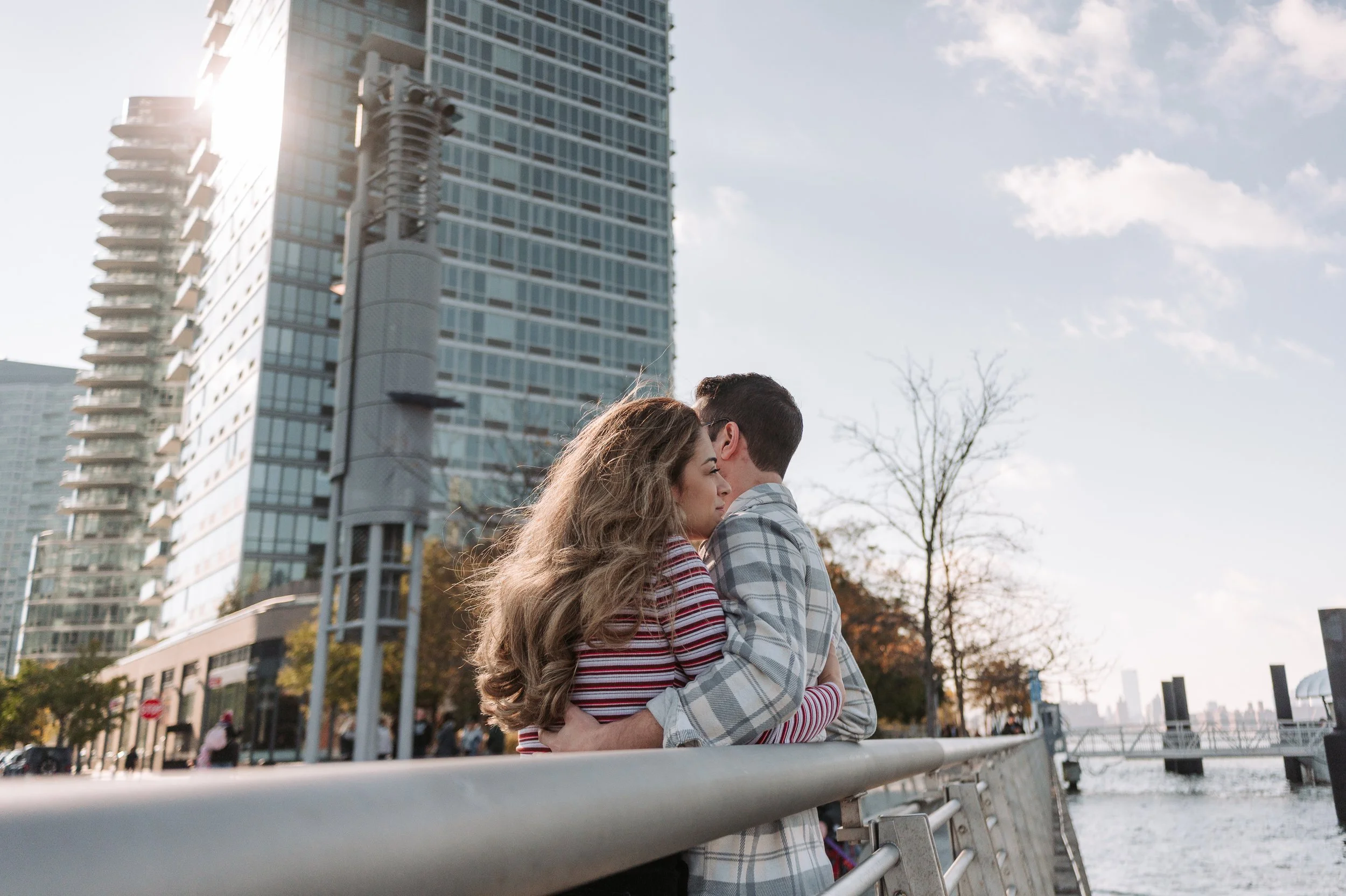 Dos personas abrazadas junto al río en una ciudad moderna, con edificios altos y árboles sin hojas en el fondo, en un día soleado.
