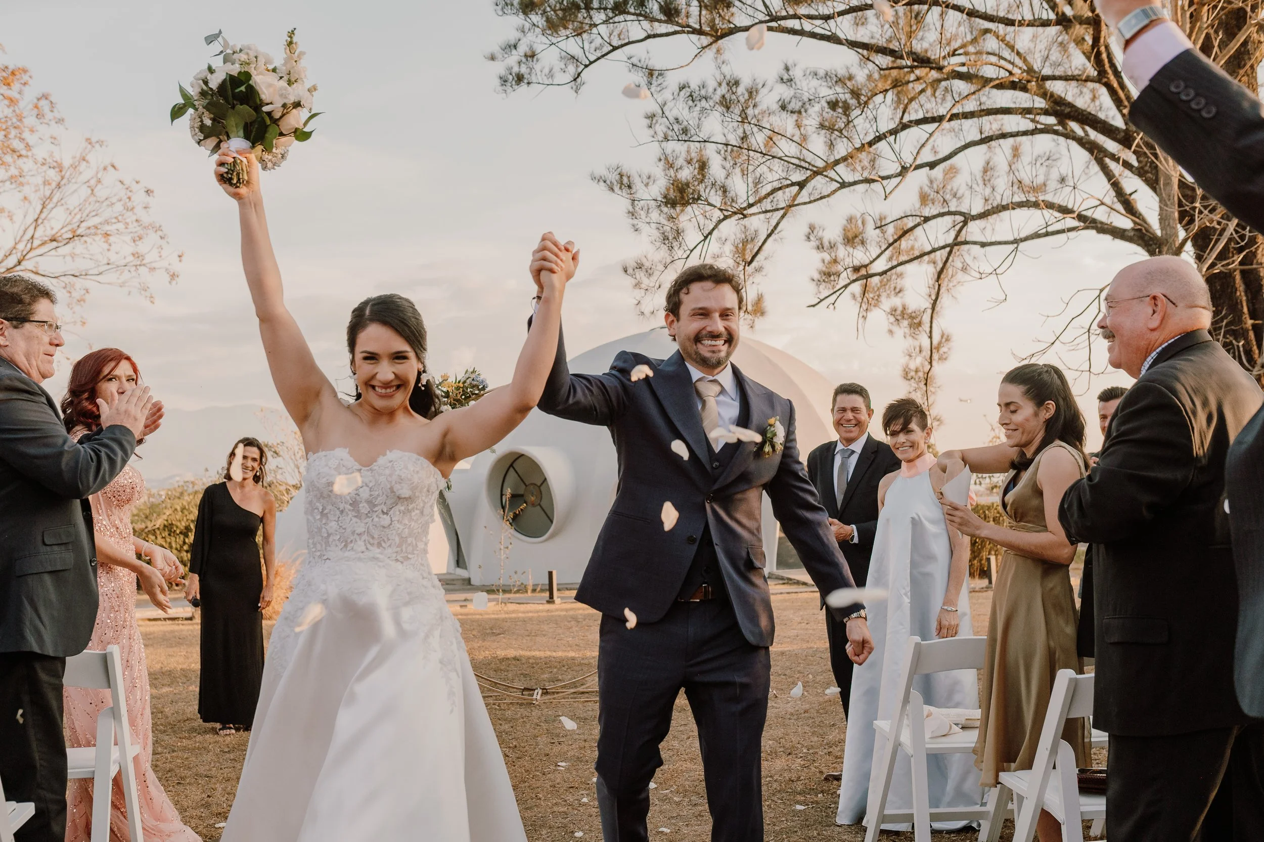 Pareja de recién casados sonriendo y celebrando en una boda al aire libre, rodeados de familiares y amigos. La novia viste un vestido de encaje y sostiene ramo, el novio lleva traje oscuro y corbata. La escena tiene árboles y cielo de fondo durante atardecer.