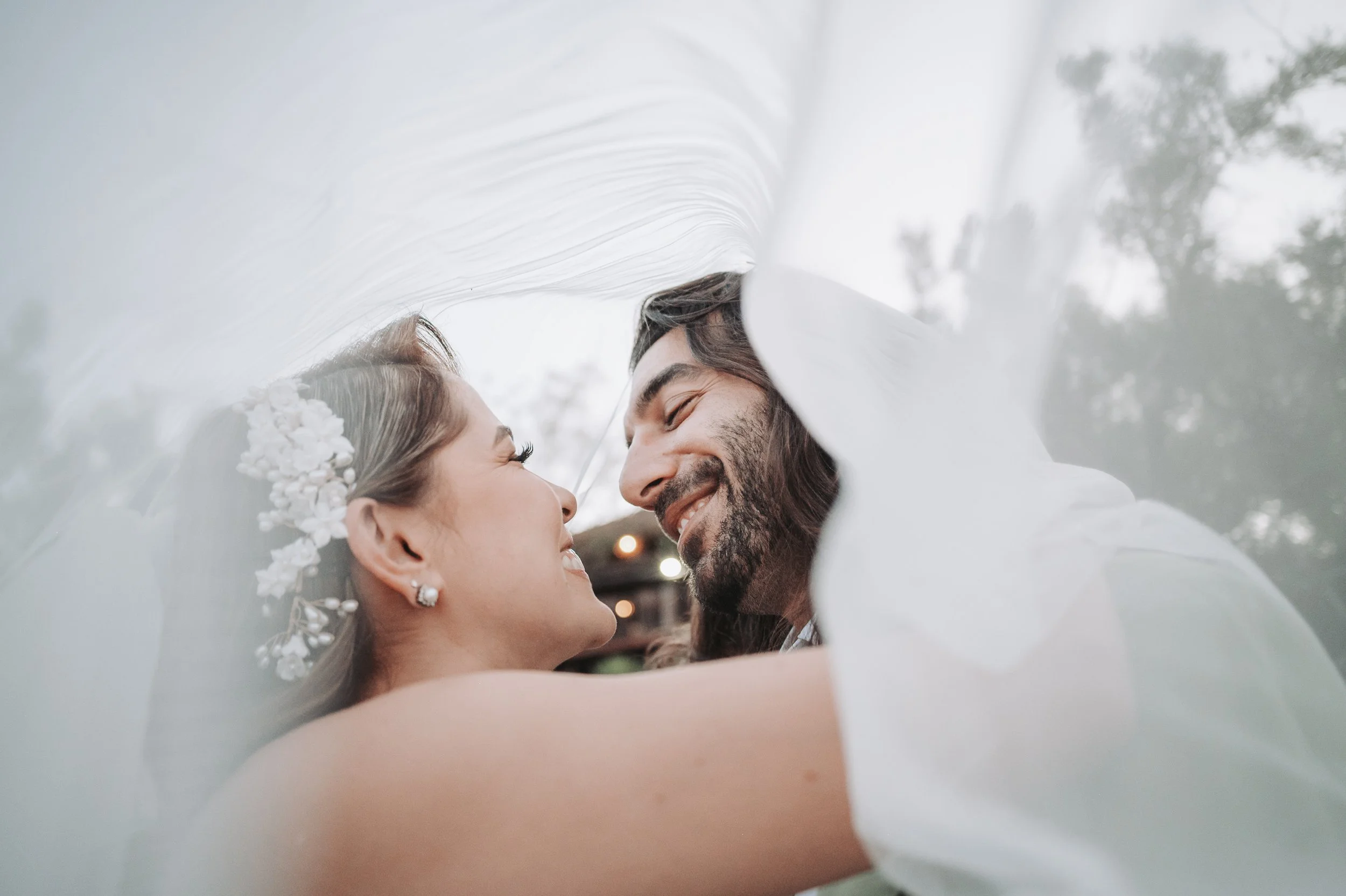 Pareja de recién casados sonriendo y mirándose con una tela de novia sobre ellos en un ambiente exterior.