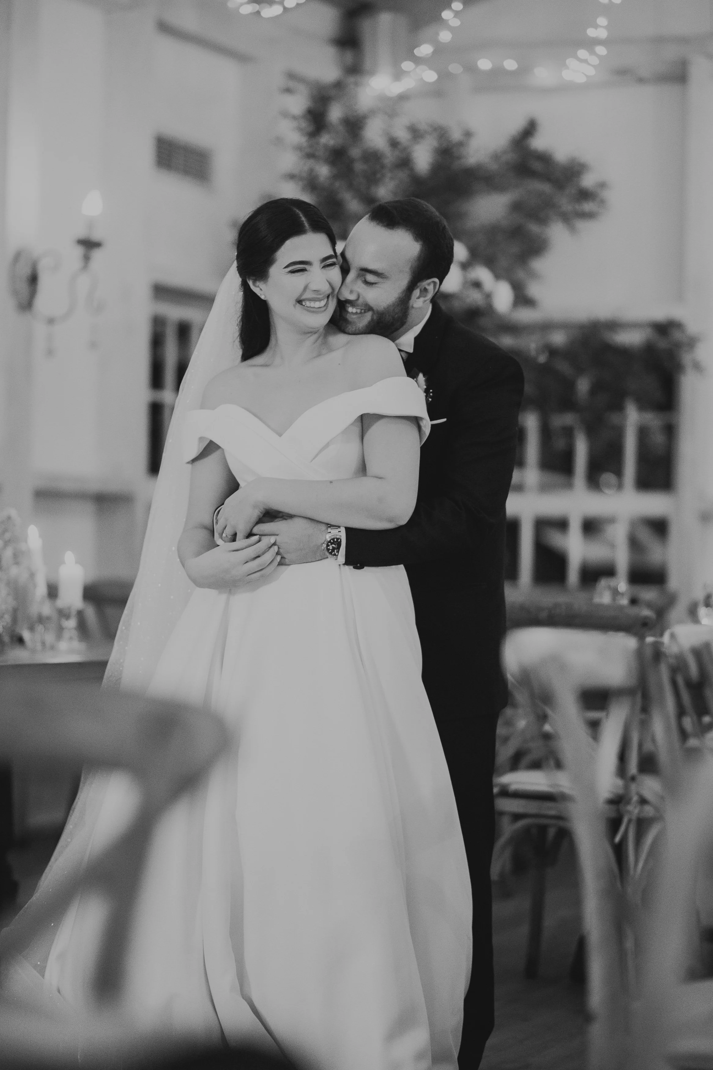 Una pareja de recién casados sonriendo y abrazándose en una celebración de boda, en un ambiente interior decorado con luces y velas.