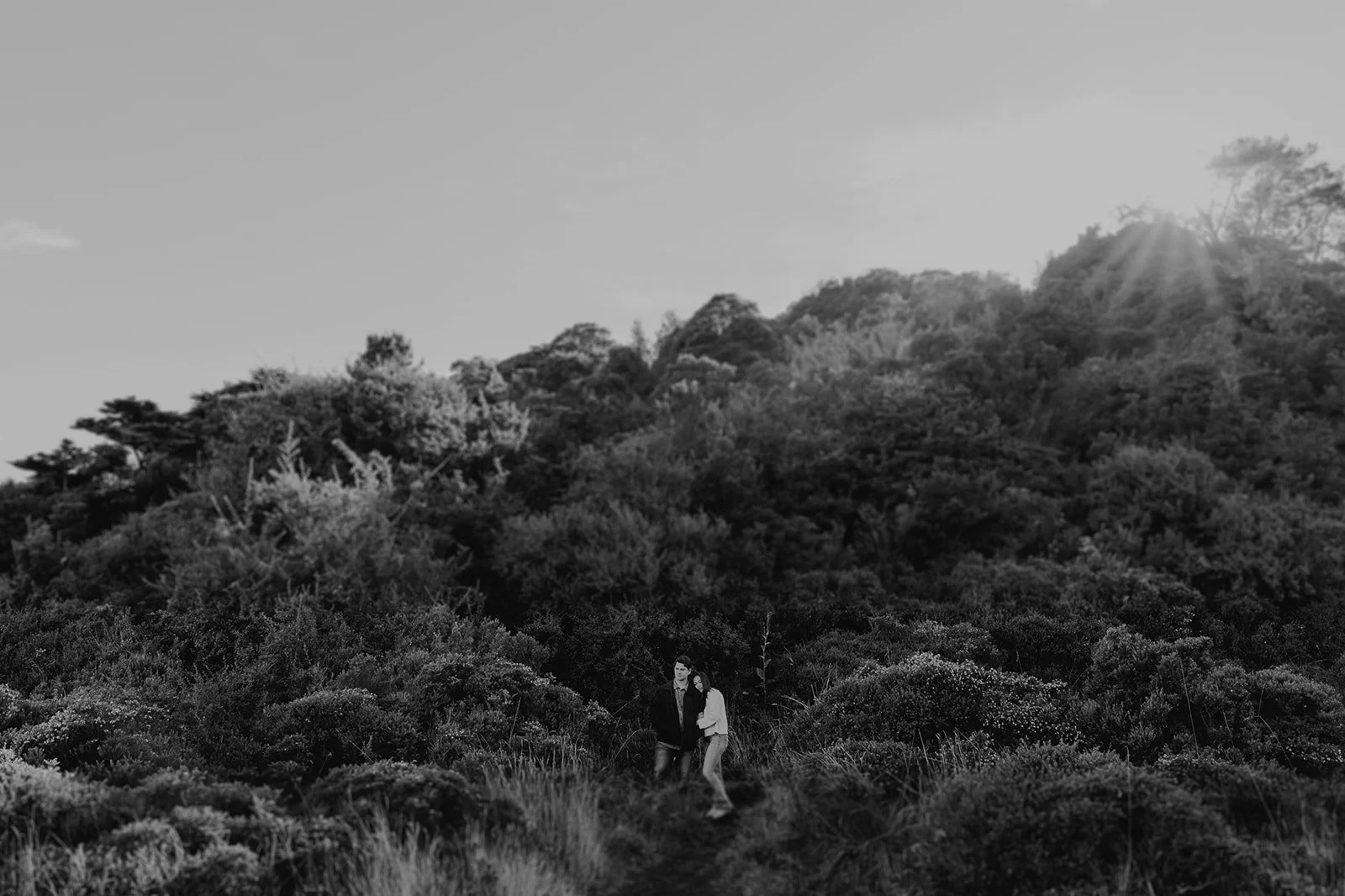 Una pareja de pie en medio de un campo con vegetación y árboles frondosos, con el sol brillando sobre los árboles al fondo.
