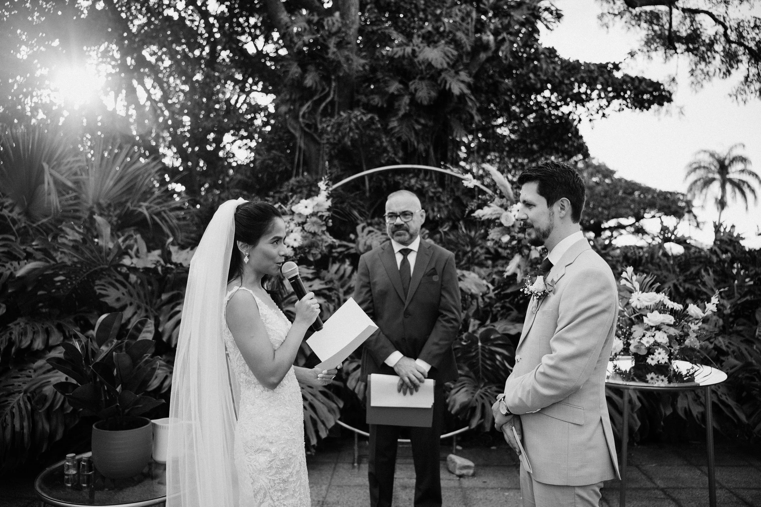 Una pareja de novios en su ceremonia de boda, la novia sostiene un micrófono y lee votos, mientras que el novio la mira sonriendo. Un oficiante está en el centro, con un fondo de plantas tropicales y decoración floral.