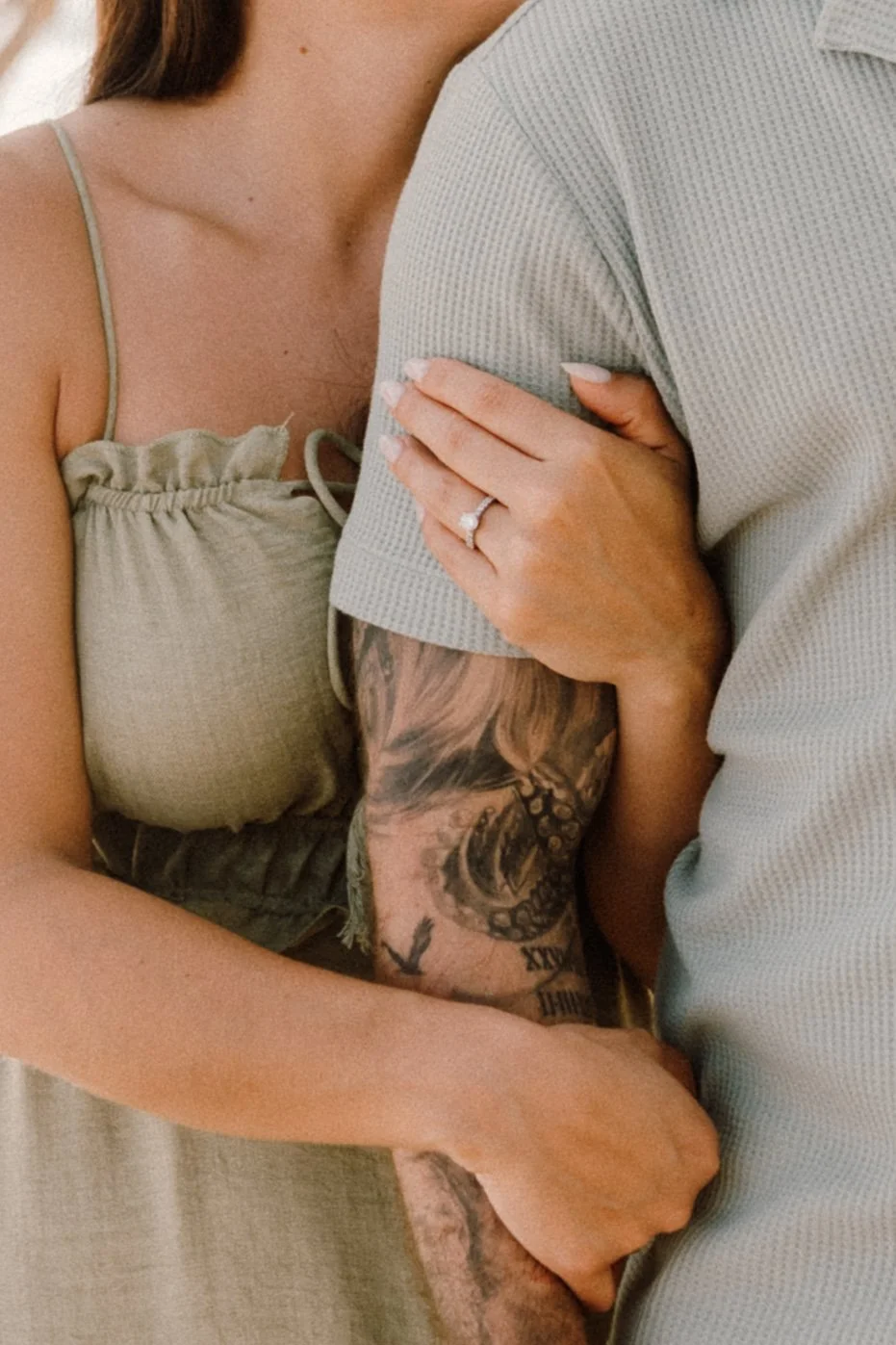 Una pareja abrazándose, mostrando un anillo de compromiso en la mano de la mujer, con tatuajes en el brazo del hombre.