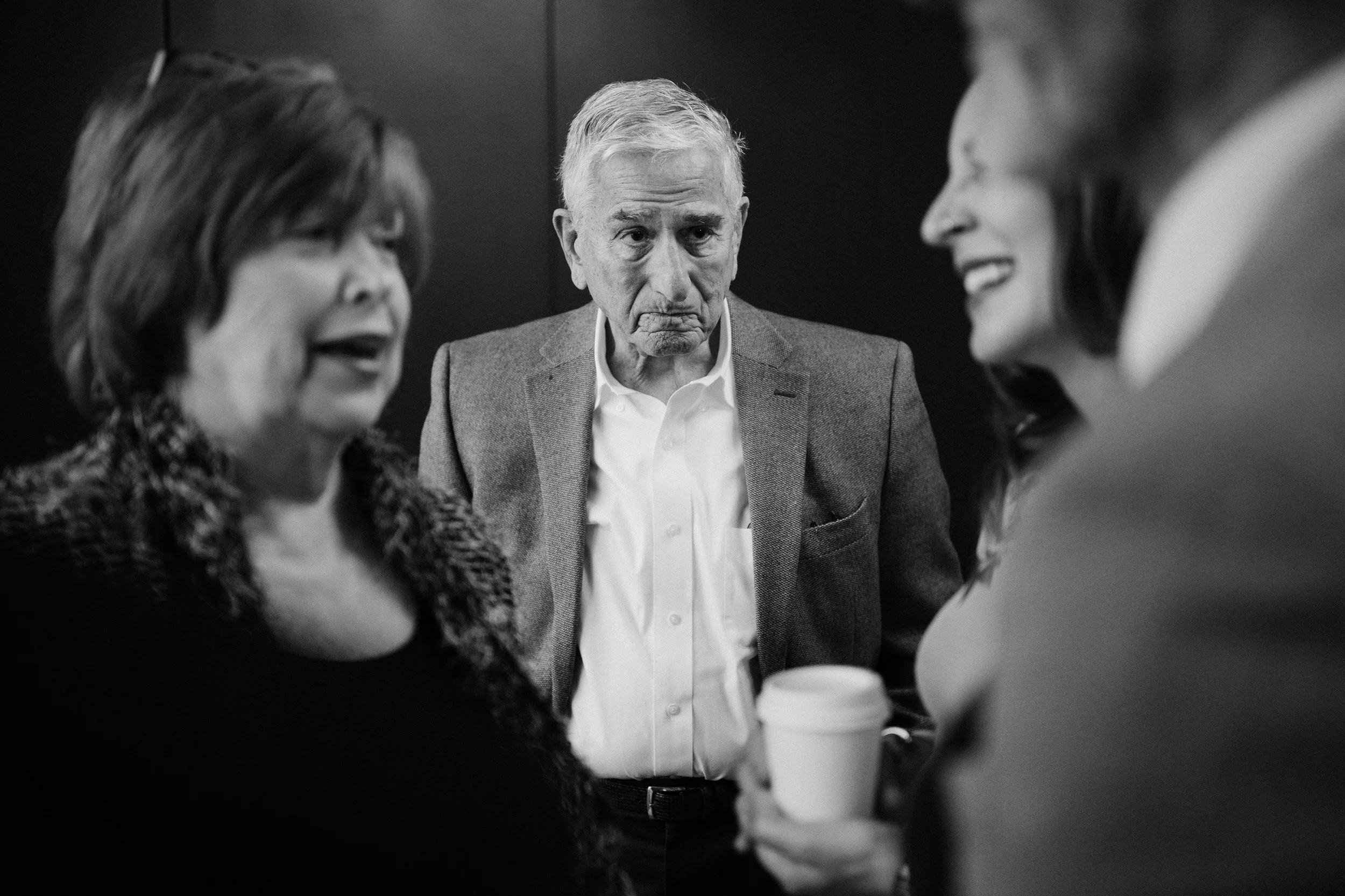 Tres personas, dos mujeres y un hombre mayor, en una conversación en un entorno informal, con expresiones serias y una taza de café en primer plano.