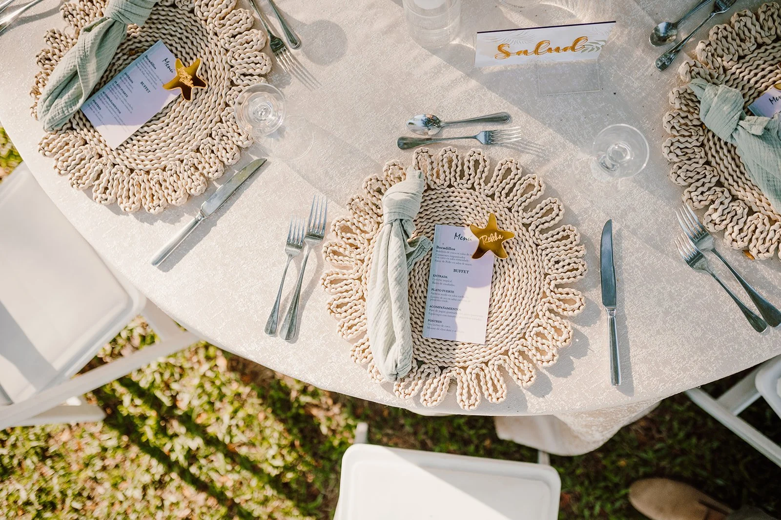 Mesa decorada con mantel blanco, platos decorativos de cuerda, cubiertos de acero inoxidable, vasos de agua, servilletas atadas con cuerda, y tarjetas con nombre, en un evento al aire libre.