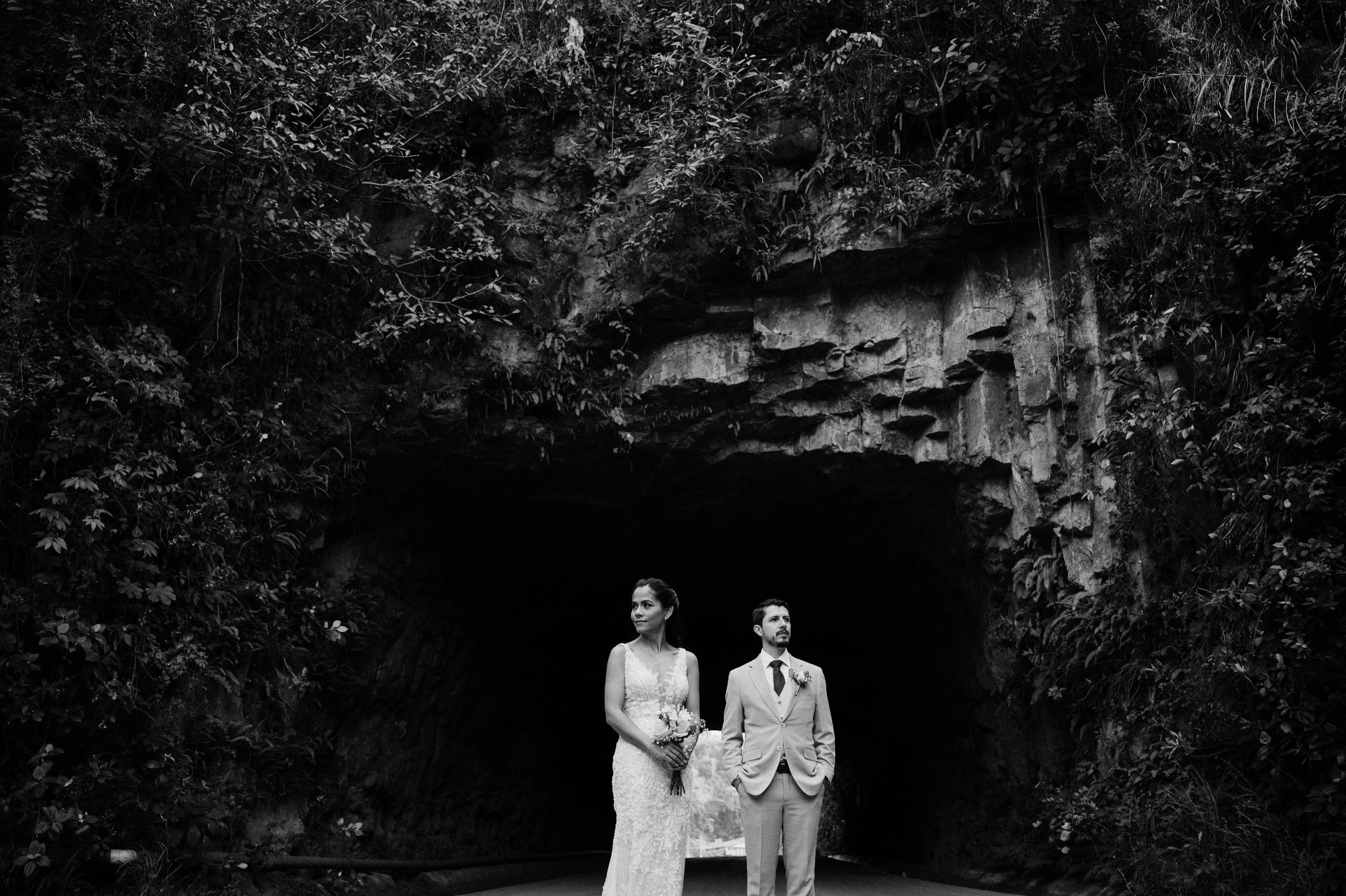 Pareja de novios en un túnel de roca, en blanco y negro, la novia con vestido de encaje y ramo de flores, el novio con saco claro y corbata.