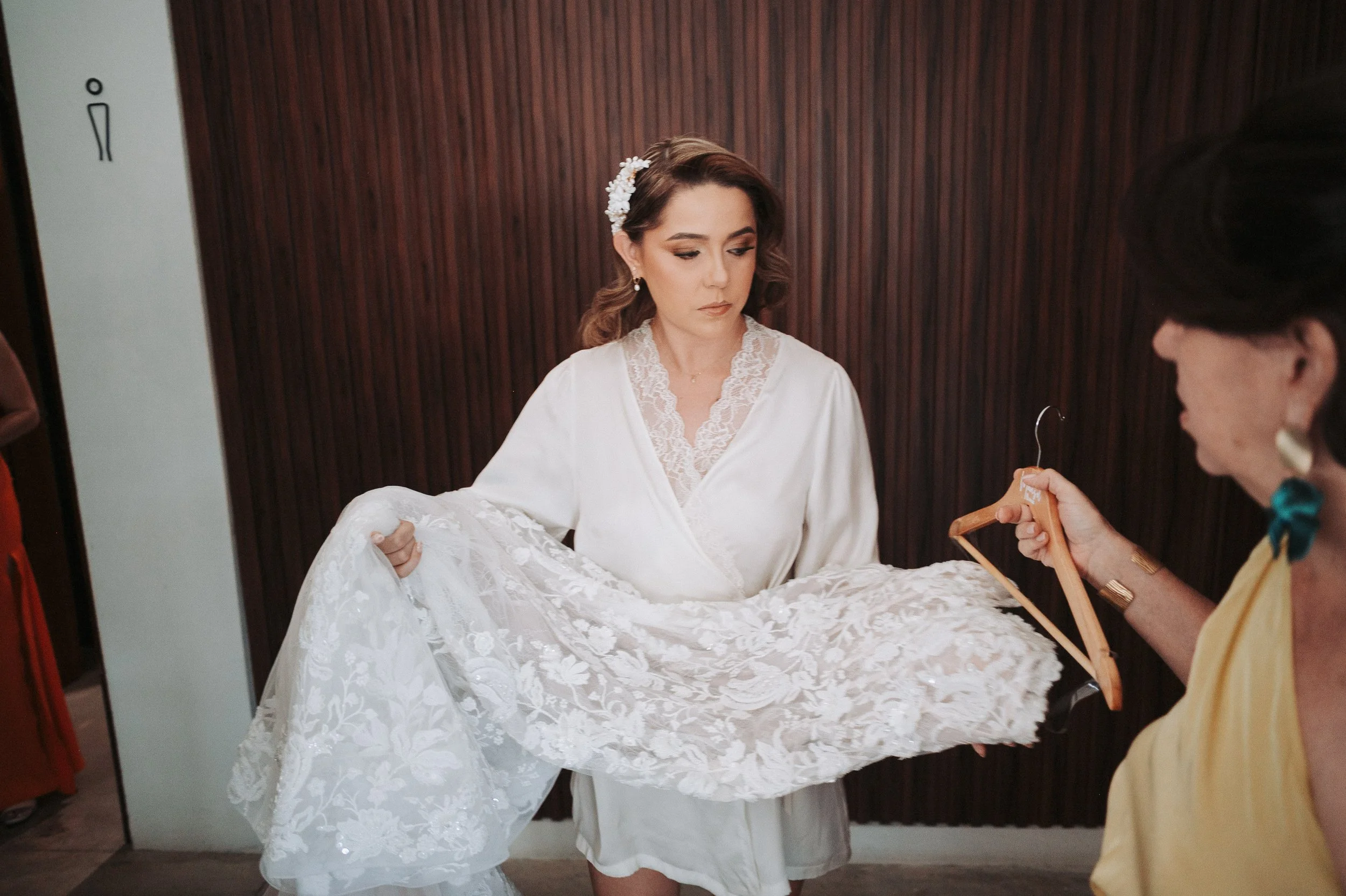 Mujer vestida con bata blanca sosteniendo un vestido de novia de encaje, mientras una persona con gafas y vestido amarillo le toma una foto con un teléfono móvil en un lugar con paredes de madera.