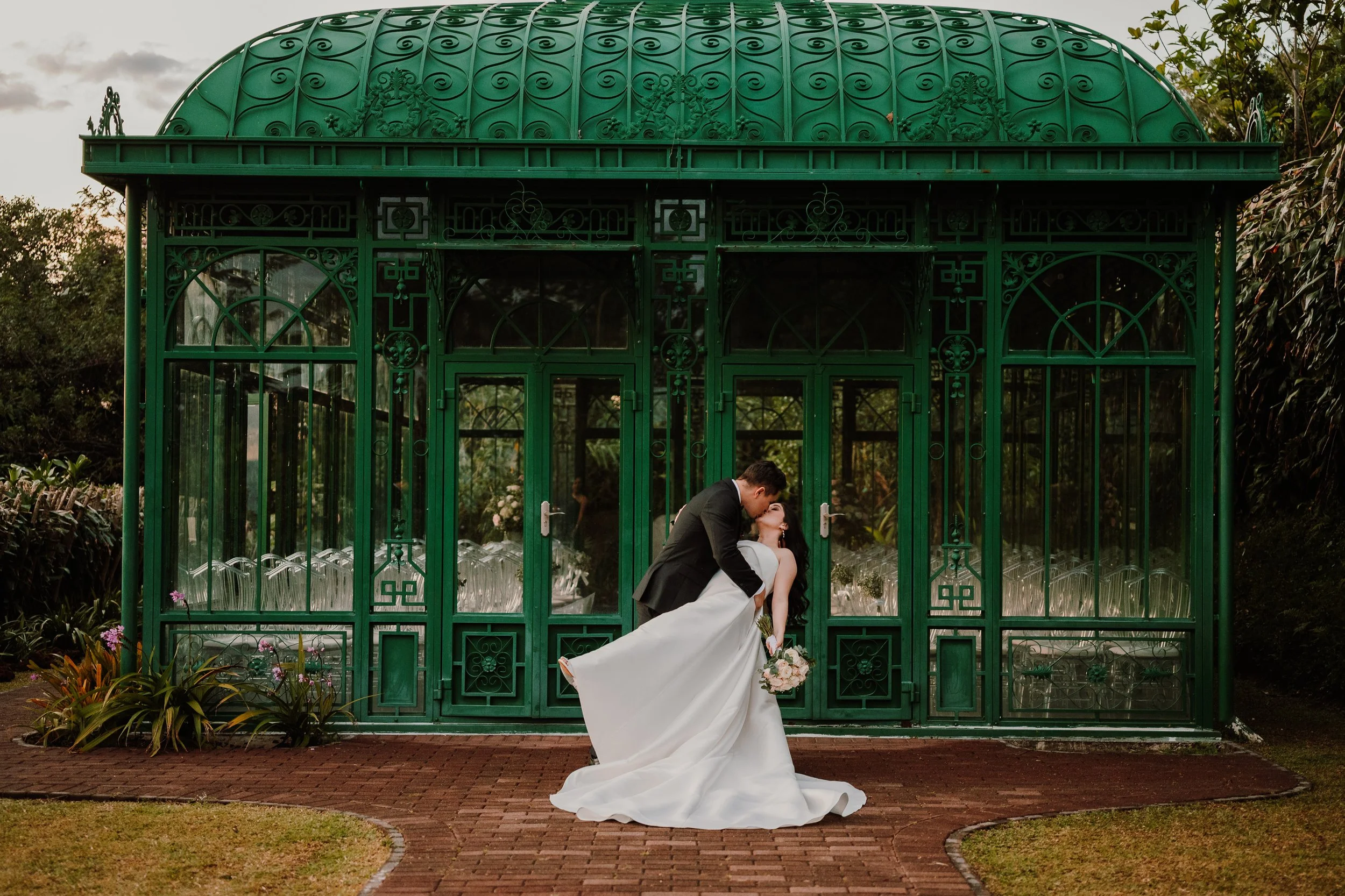 Pareja de recién casados besándose en un invernadero verde en un parque durante el atardecer.