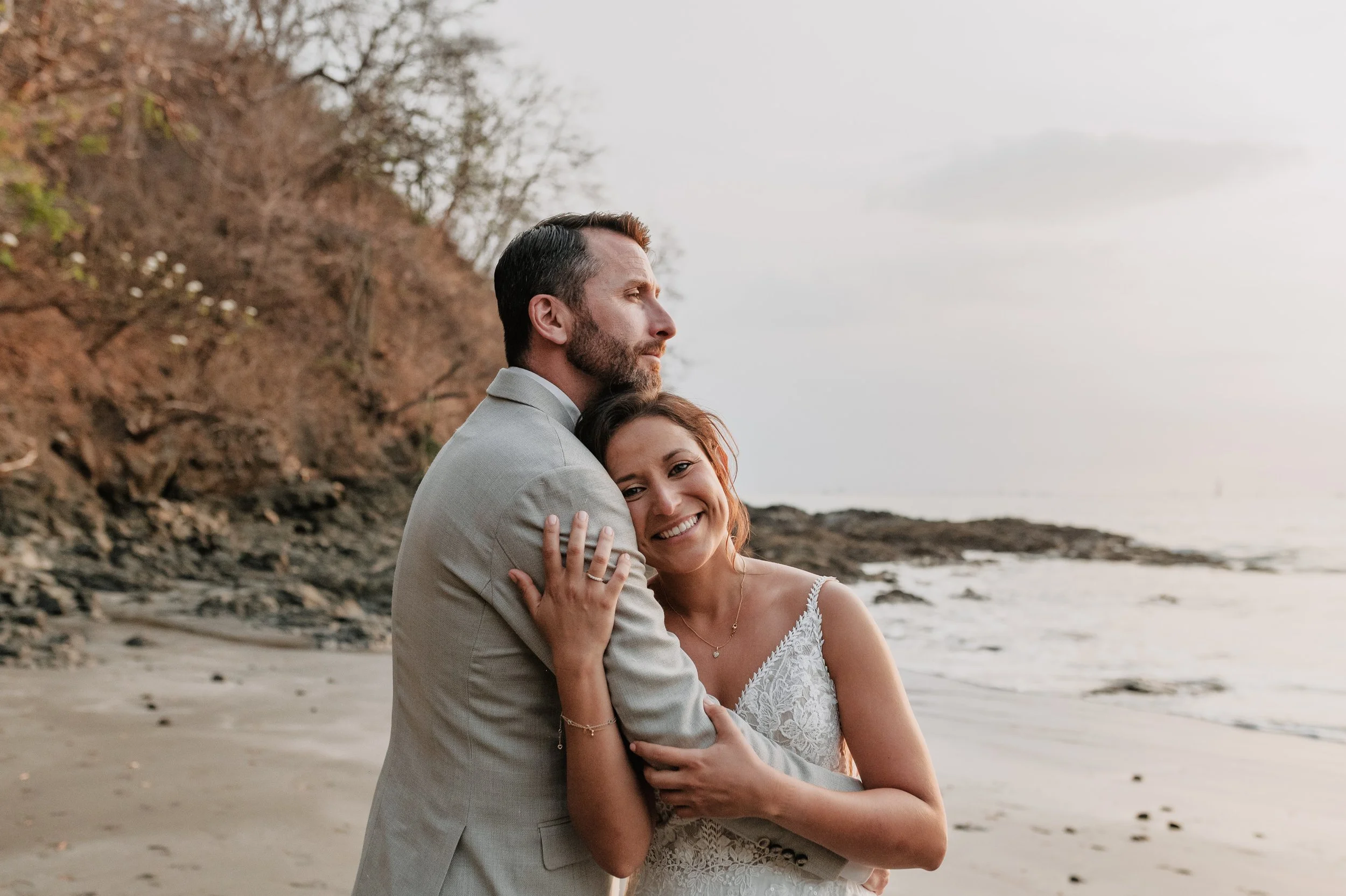 Pareja feliz en la playa, abrazados, disfrutando de la vista del mar y el atardecer, con árboles y rocas en el fondo.