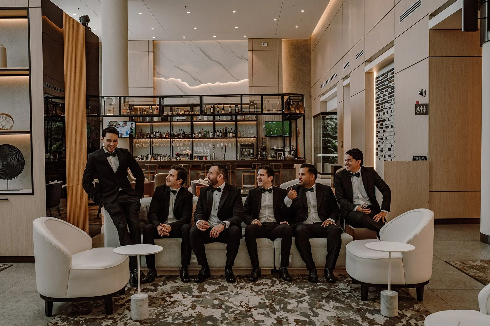 Grupo de siete hombres vestiendo trajes formales en un salón elegante, sonriendo y conversando, con un bar visible en el fondo.