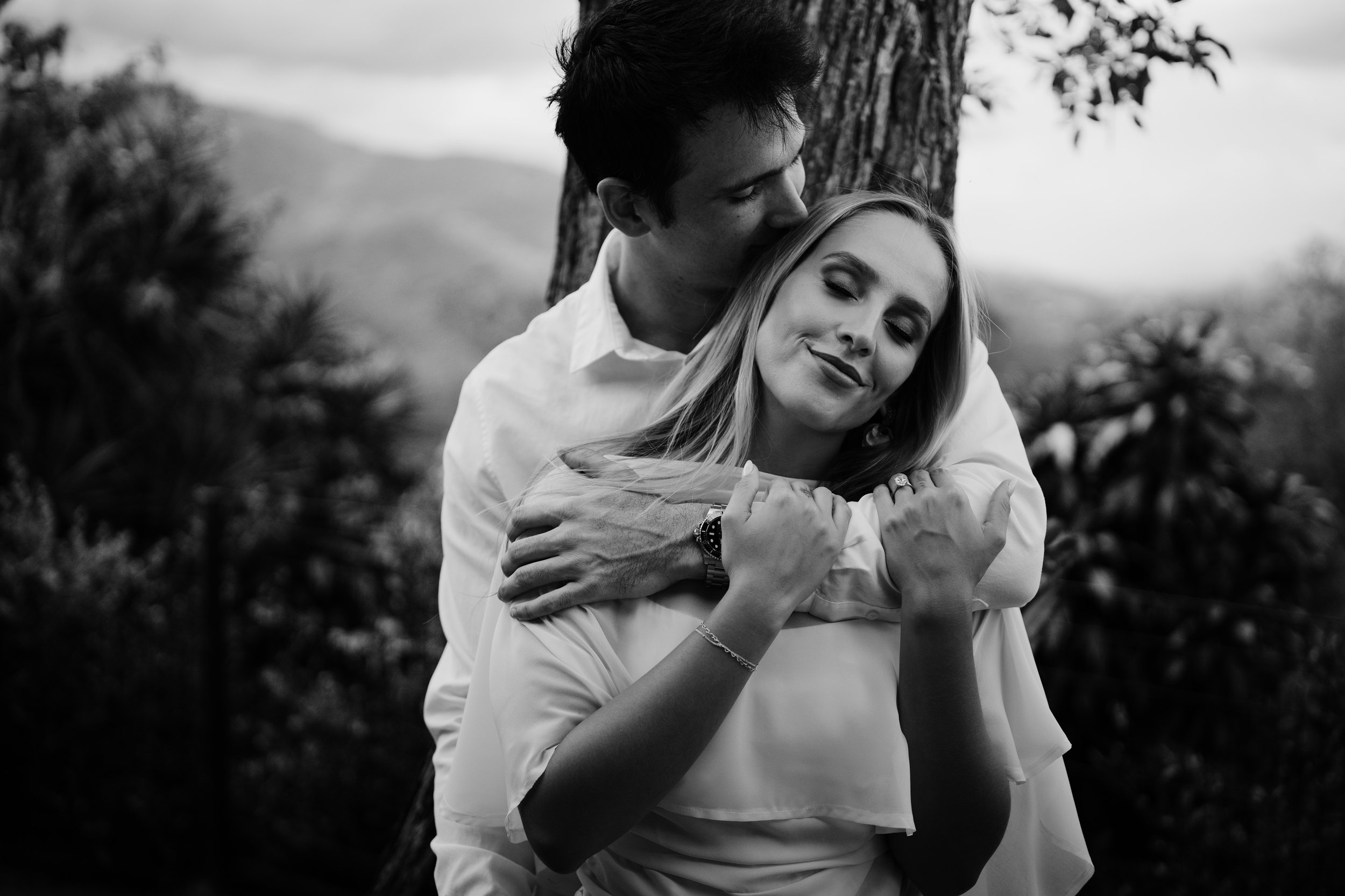 Una pareja abrazada y sonriendo en un entorno natural, ella con ojos cerrados y él besándola en la frente, en blanco y negro.