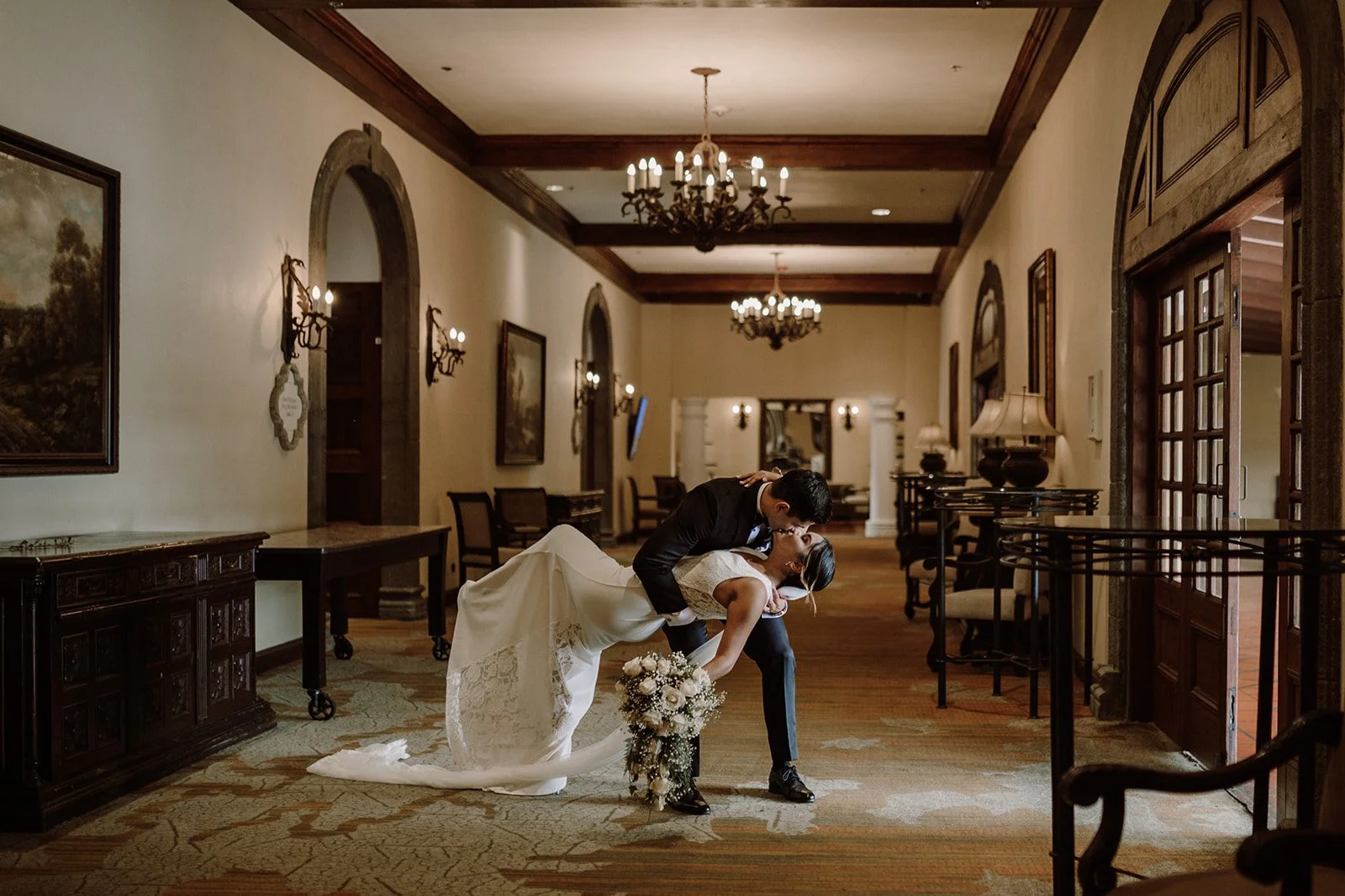 Pareja de novios en un salón elegante, el novio sostiene a la novia en una pose romántica, la novia lleva un vestido blanco y sostiene un ramo de flores.