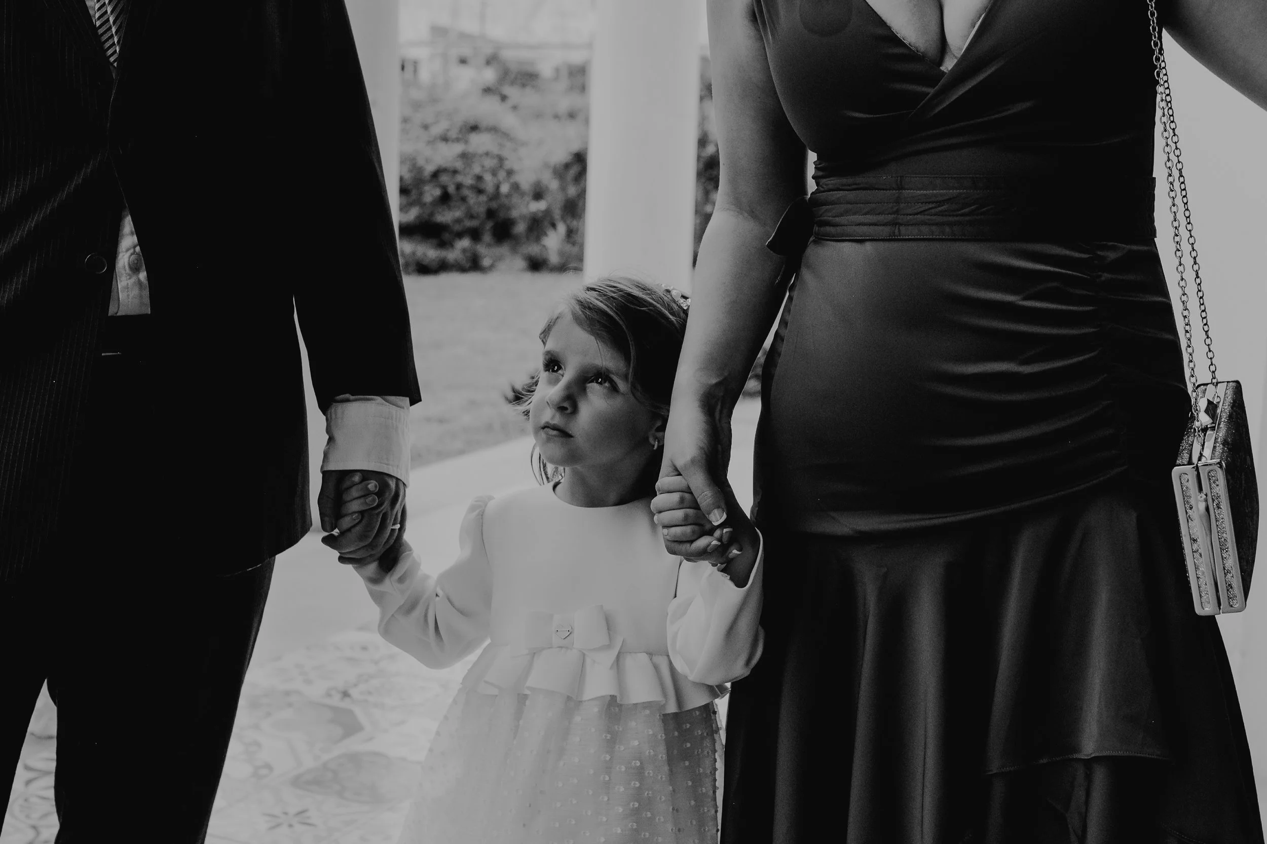 Niña con vestido blanco observa a adultos en un evento formal, sosteniendo de las manos a un hombre y una mujer