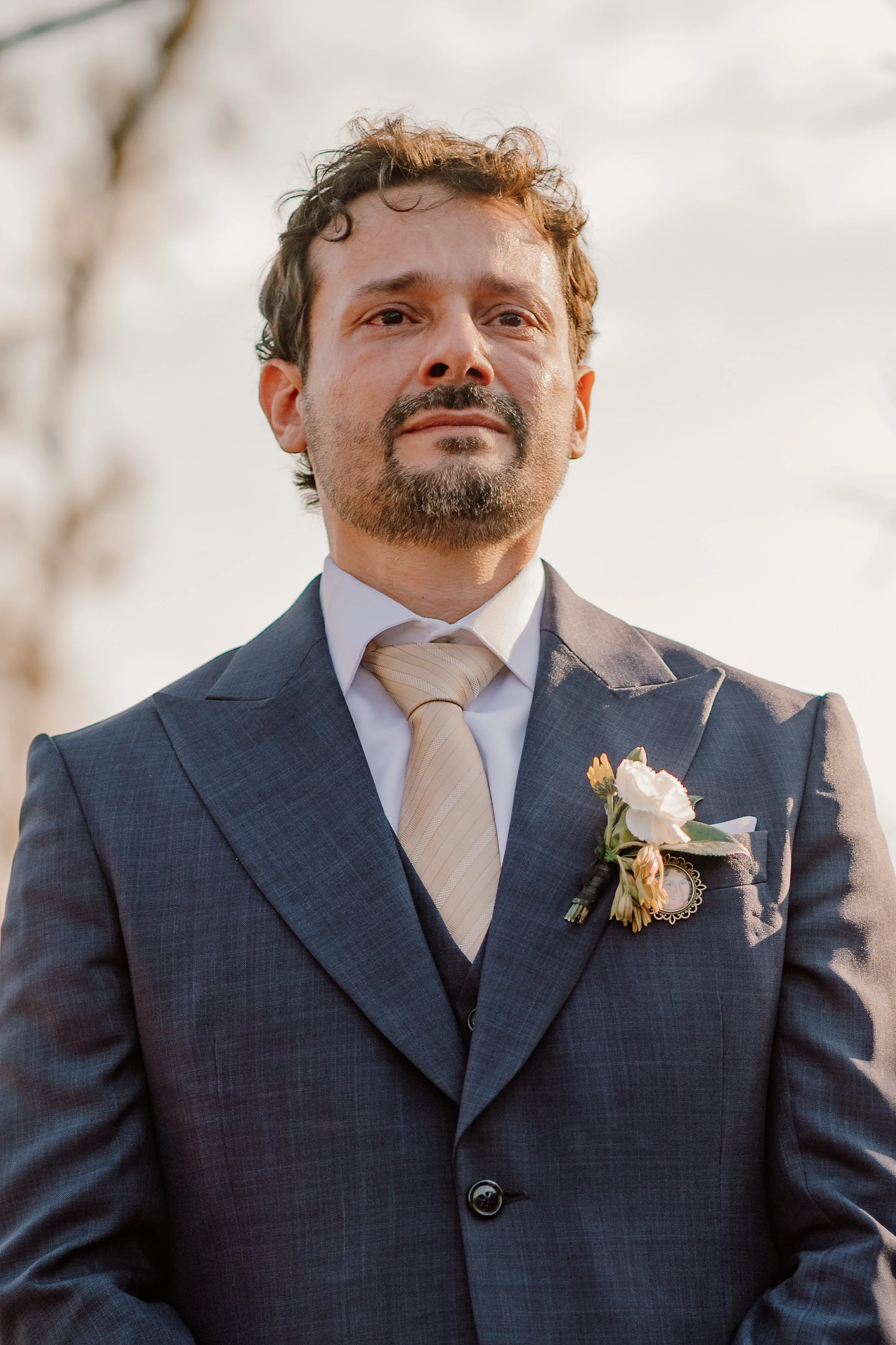 Hombre con barba y bigote, vestido con traje gris, camisa blanca y corbata crema, con flor en la solapa, en un fondo de cielo y árboles.