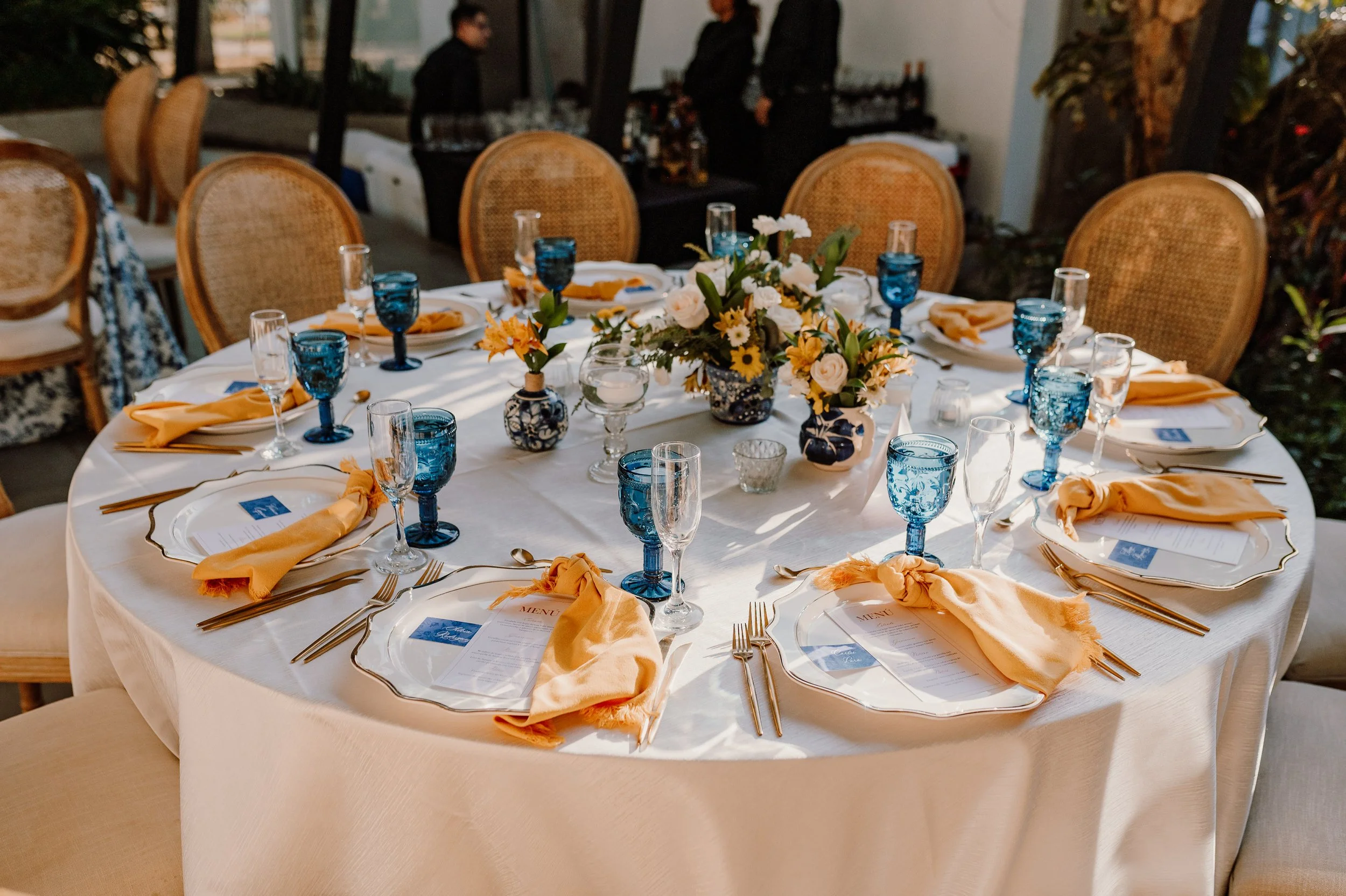 Mesa decorada para evento con flores en el centro, platos, copas y cubiertos, servilletas naranja, en un ambiente elegante y bien iluminado.