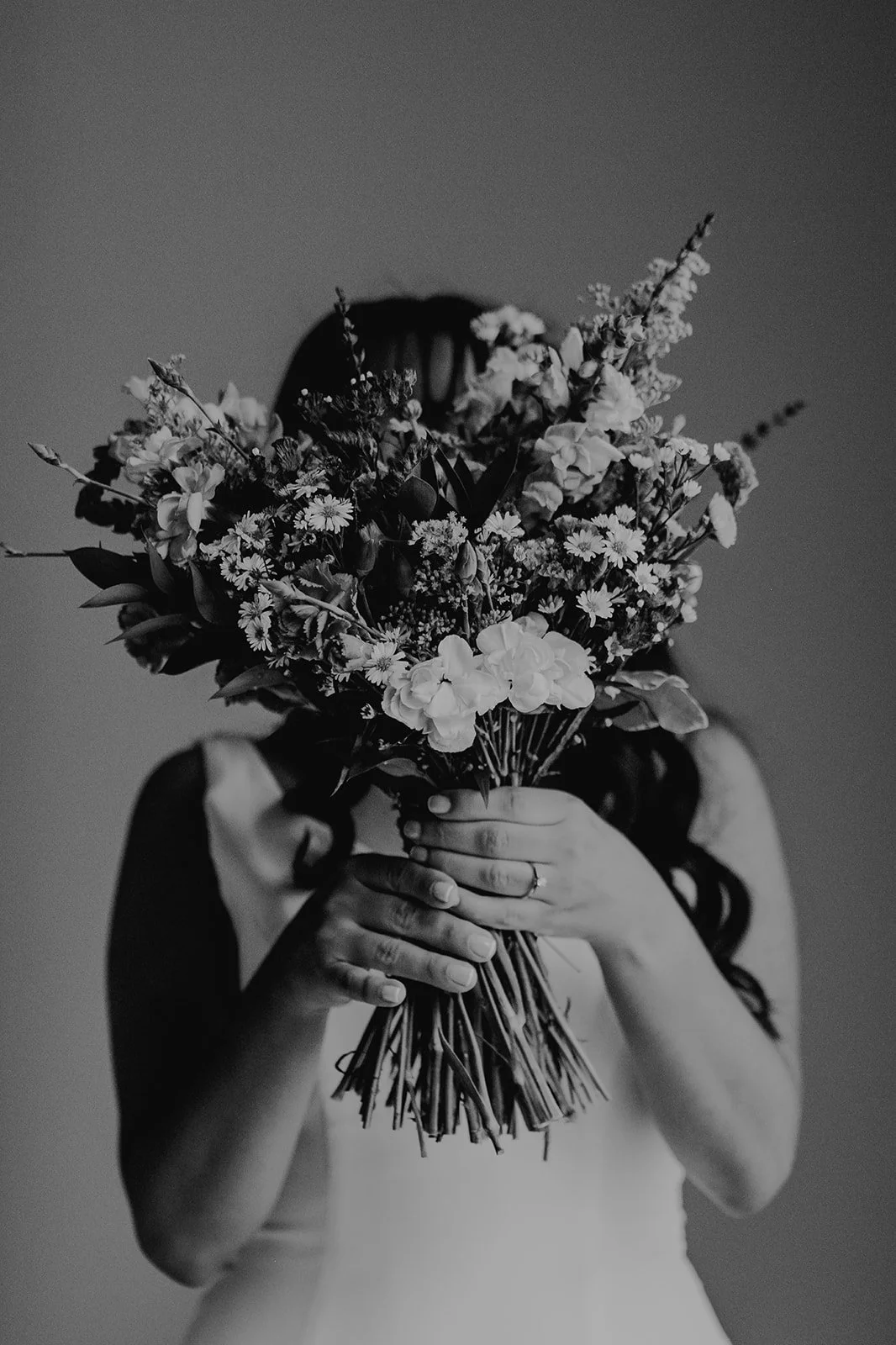 Una persona con vestido blanco sosteniendo un ramo de flores con la cara cubierta por las flores, en una foto en blanco y negro.