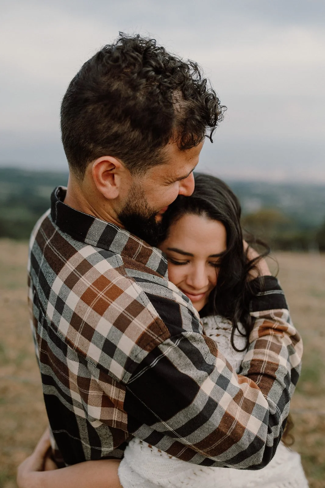 Una pareja abraza con cariño en un campo, con un fondo de cielo nublado.