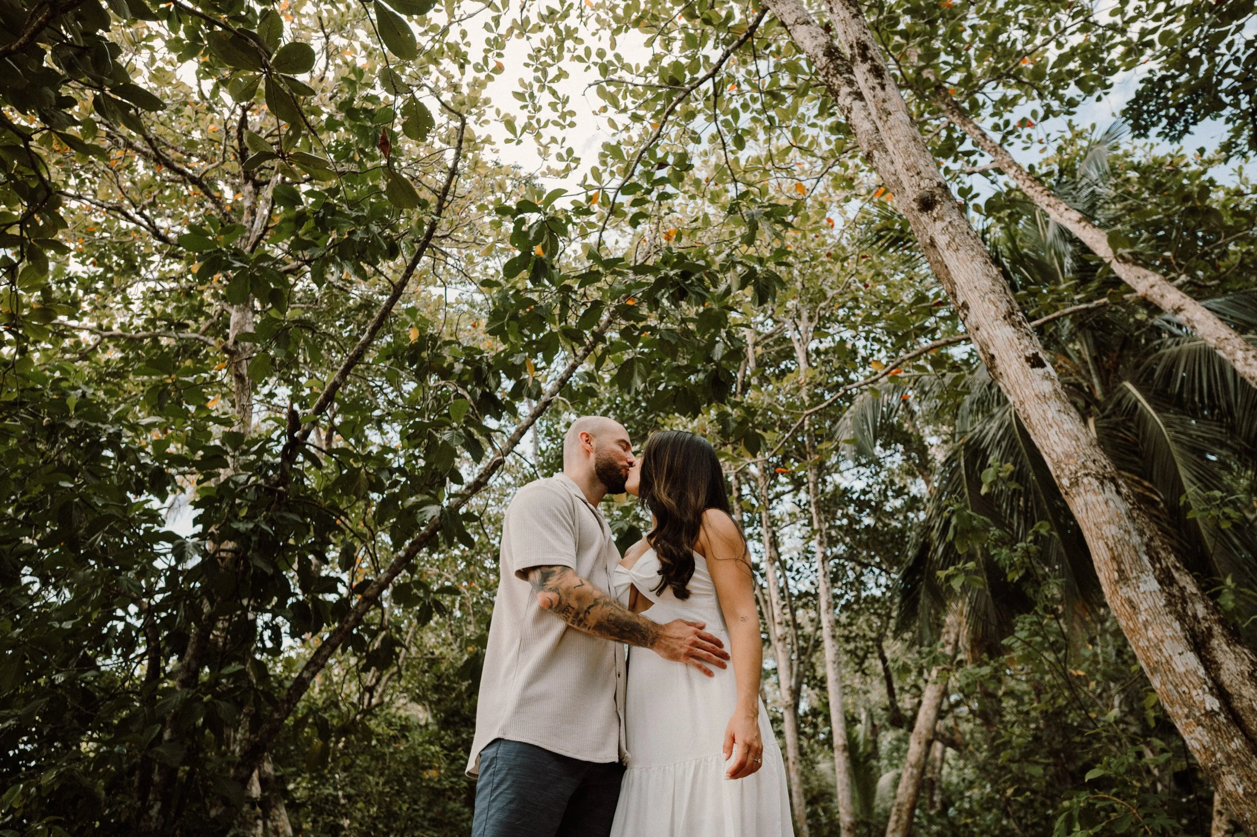 Una pareja se besa y se abraza en un entorno natural con árboles y hojas verdes.