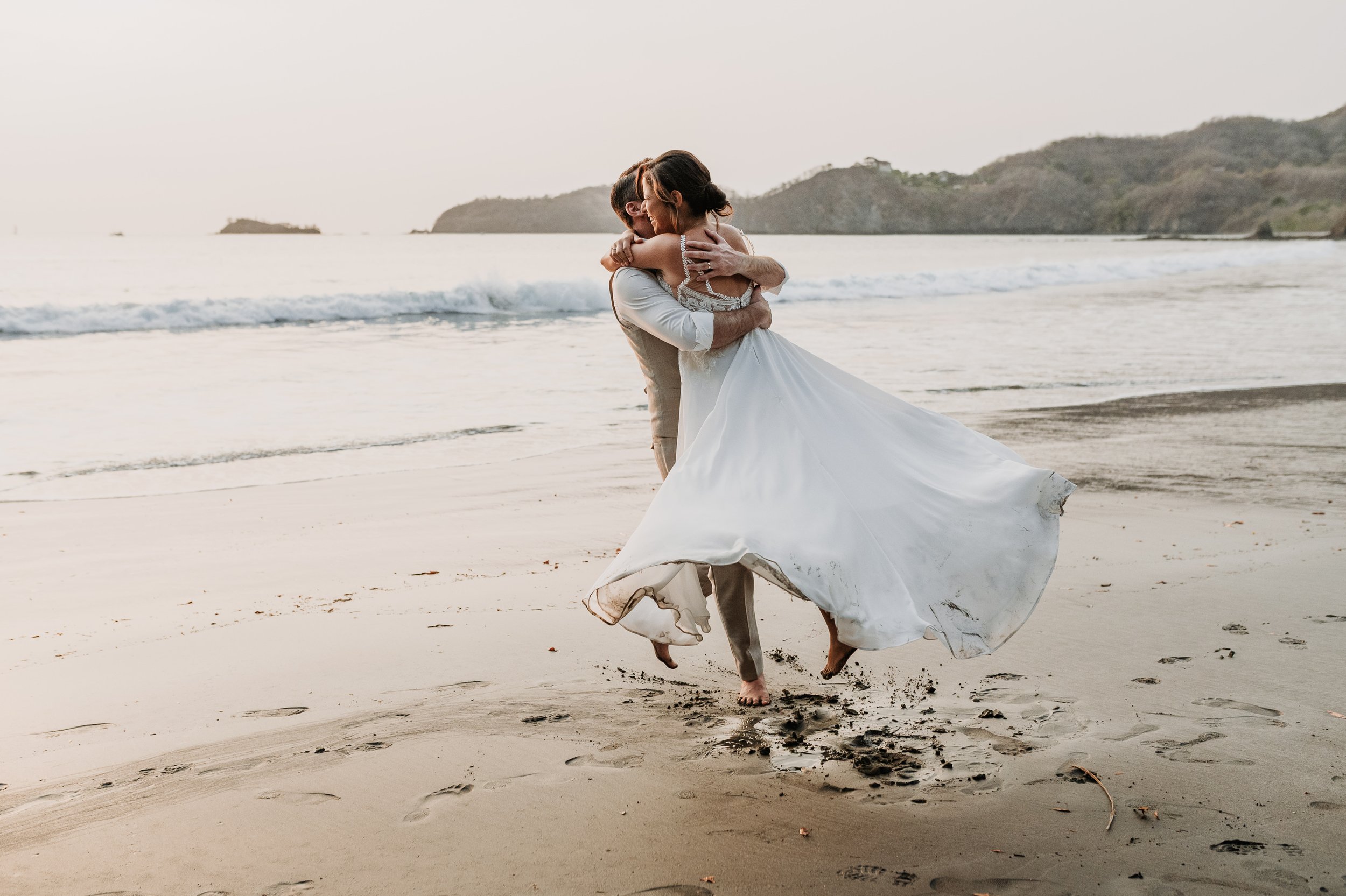 Una pareja bailando en la playa, el hombre levantando a la mujer en sus brazos, durante el atardecer.