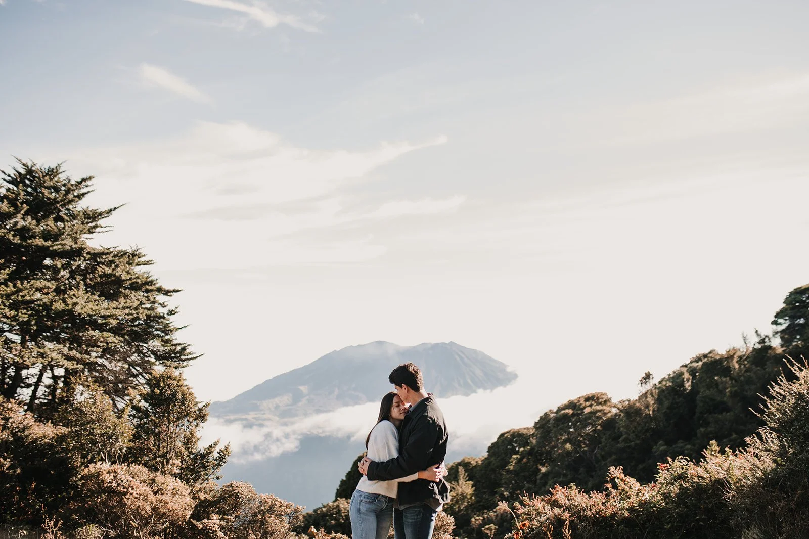 Una pareja abrazándose en un paisaje montañoso con árboles y una montaña en el fondo, cielo despejado con algunas nubes.