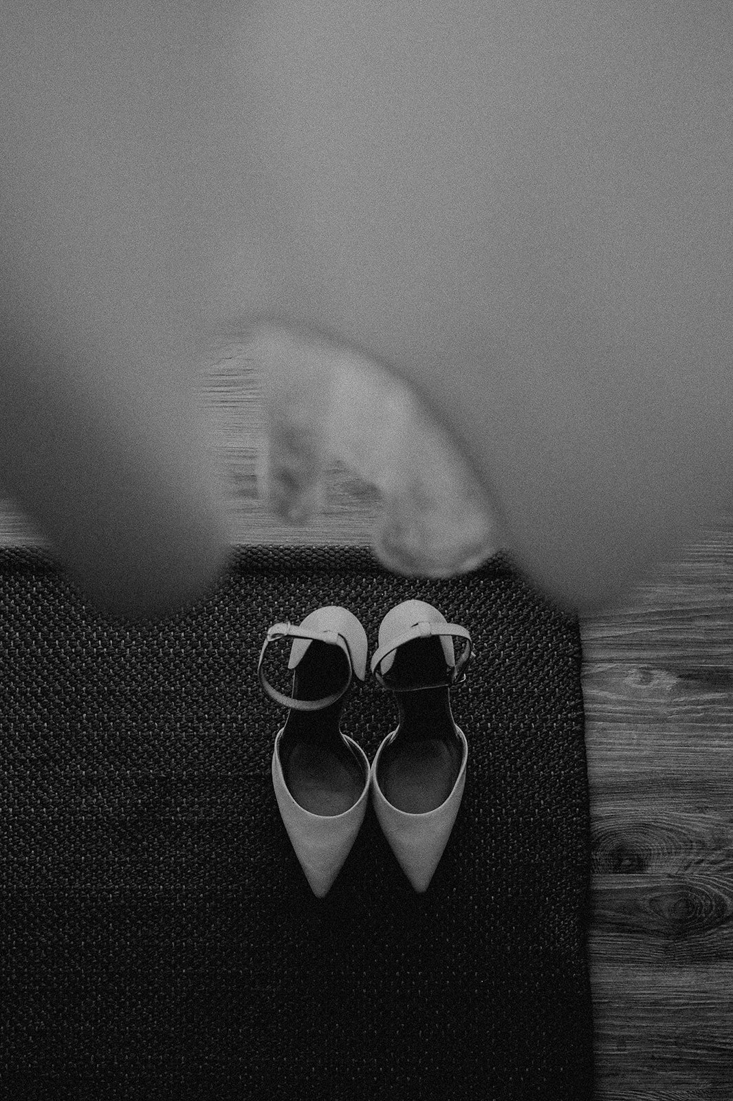 Un par de zapatos blancos de tacón en un tapete negro sobre un suelo de madera, con la sombra de una persona y un objeto en la parte superior de la imagen.