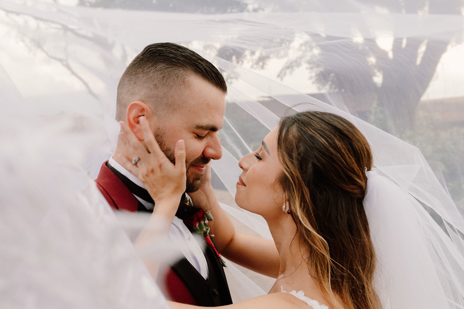 Una pareja de recién casados se mira con amor, la mujer toca la cara del hombre y ambos están sonriendo, con un velo de novia sobre ellos.