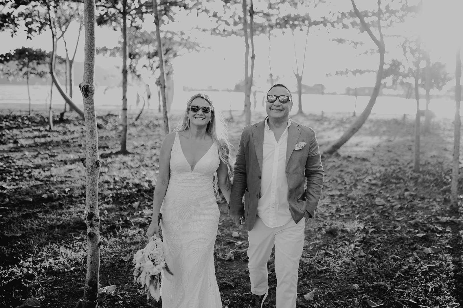 Pareja de boda caminando por un bosque, la mujer lleva un vestido blanco y sostiene un ramo, mientras que el hombre viste un saco y pantalones claros, ambos con gafas de sol.