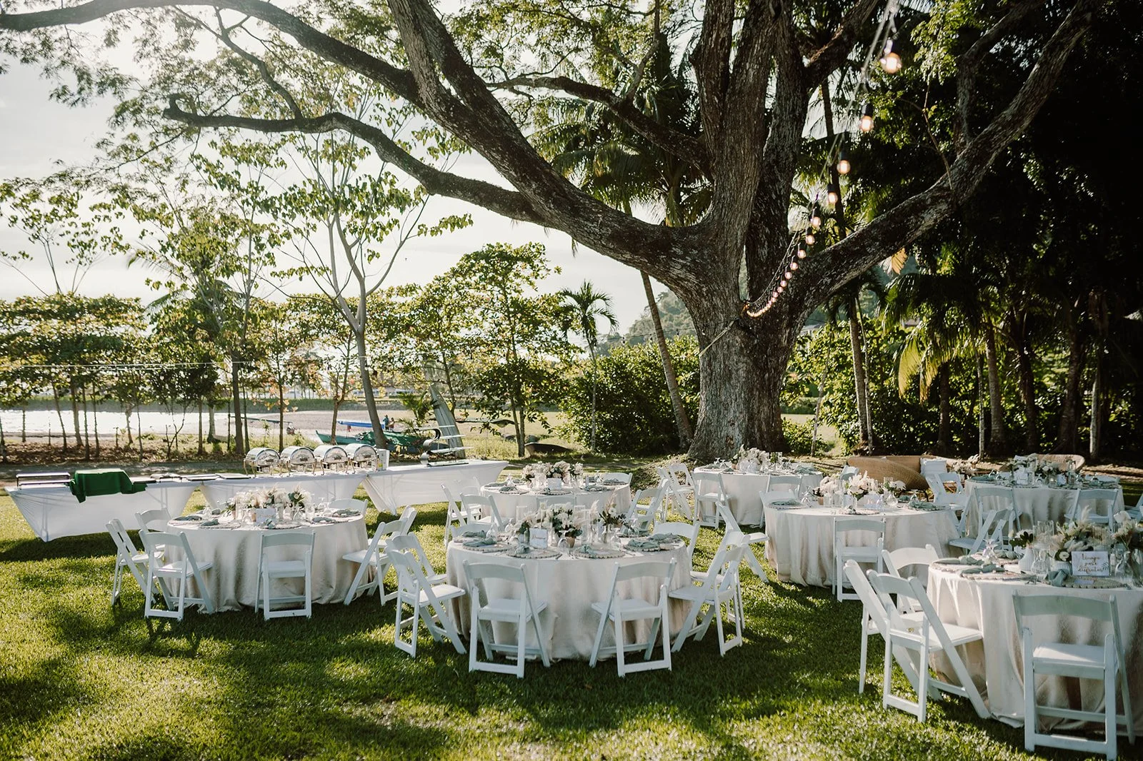 Decoración para evento al aire libre con mesas redondas con manteles blancos, sillas blancas, y adornos florales en una zona sombreada por un gran árbol. En el fondo, mesas con comida y acceso a un río o cuerpo de agua.