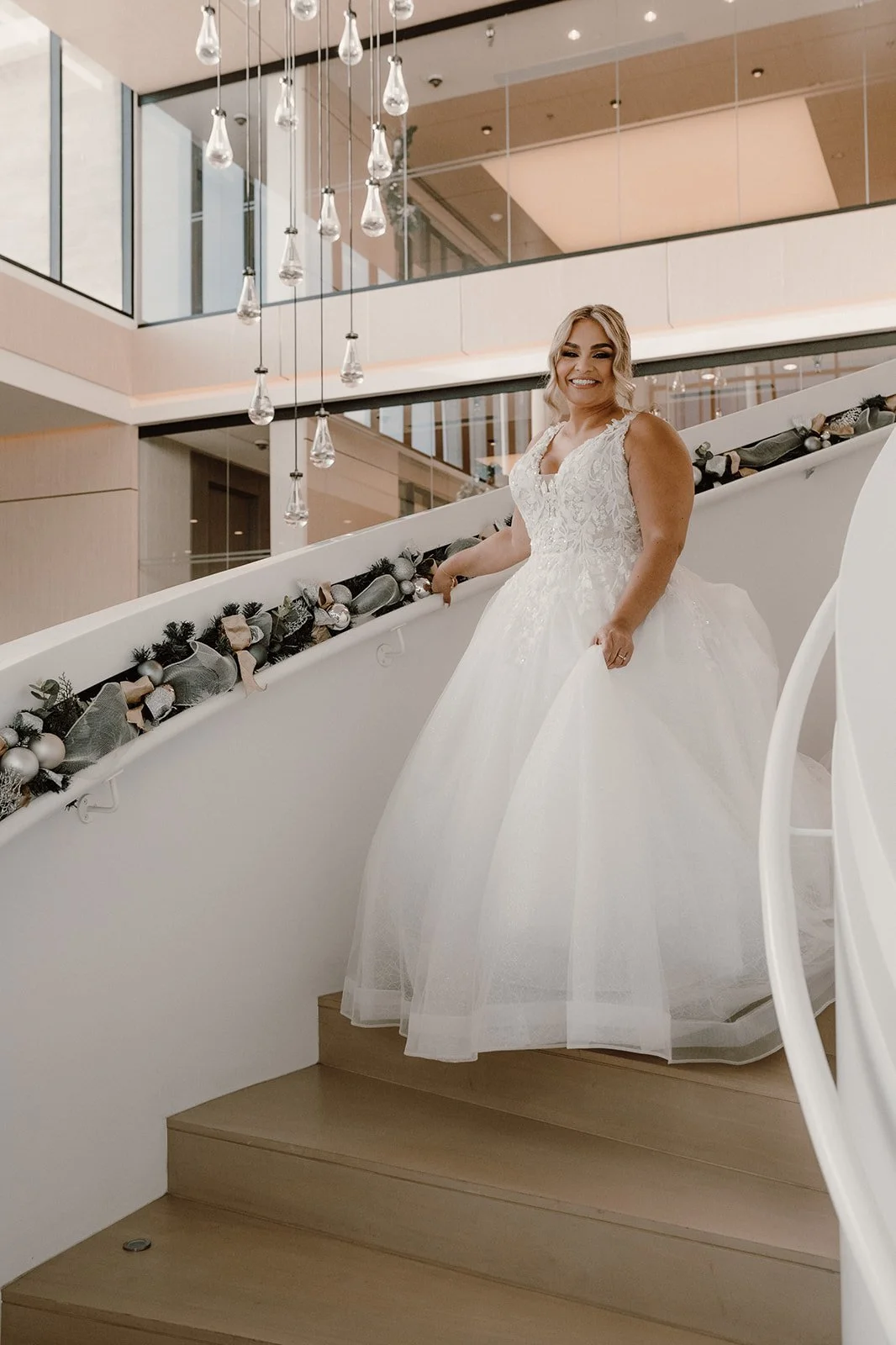 Mujer con vestido de novia en una escalinata decorada con adornos navideños, en un edificio moderno con iluminación decorativa.