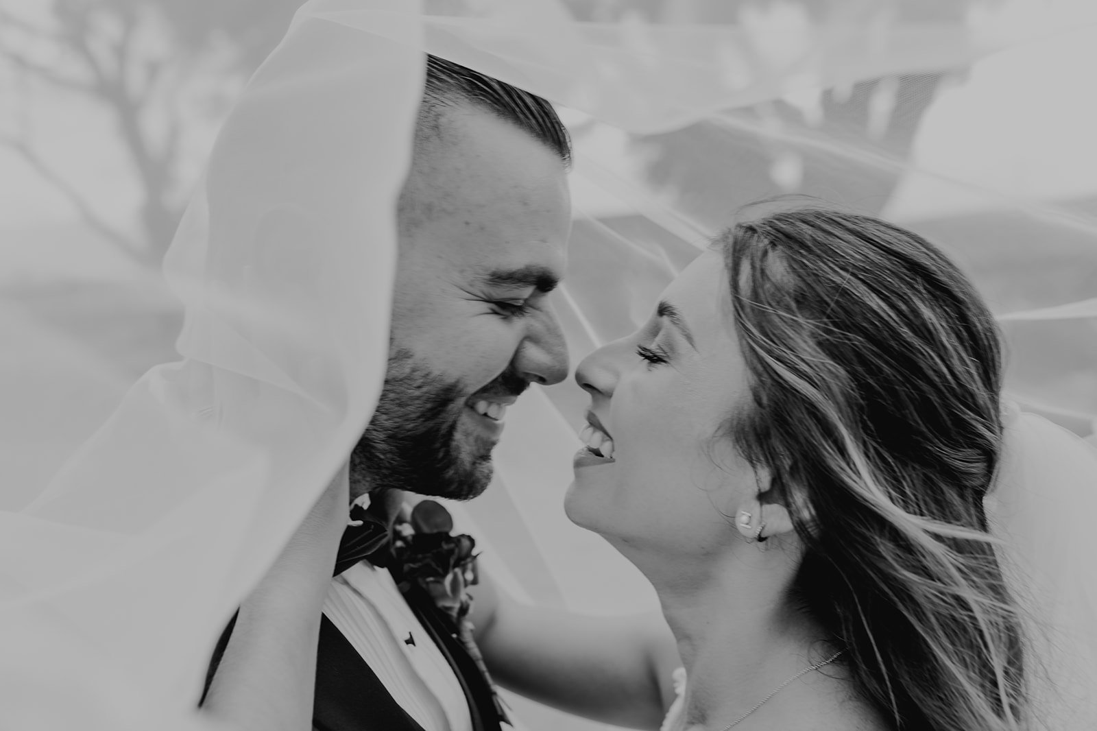 Pareja de novios en una boda, debajo de un velo nupcial, sonriendo y mirando fijamente, en blanco y negro.