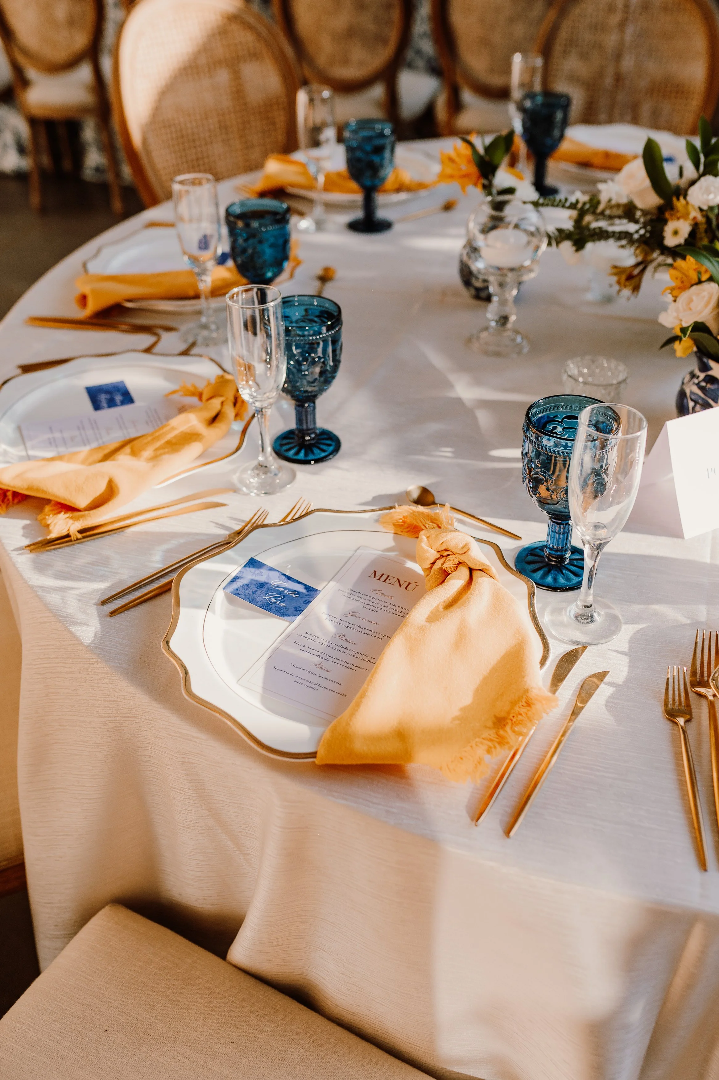 Mesa decorada para un evento con platos, cubiertos dorados, copas de cristal y vasos azules, cubiertos de tela en color durazno y un centro de mesa con flores.