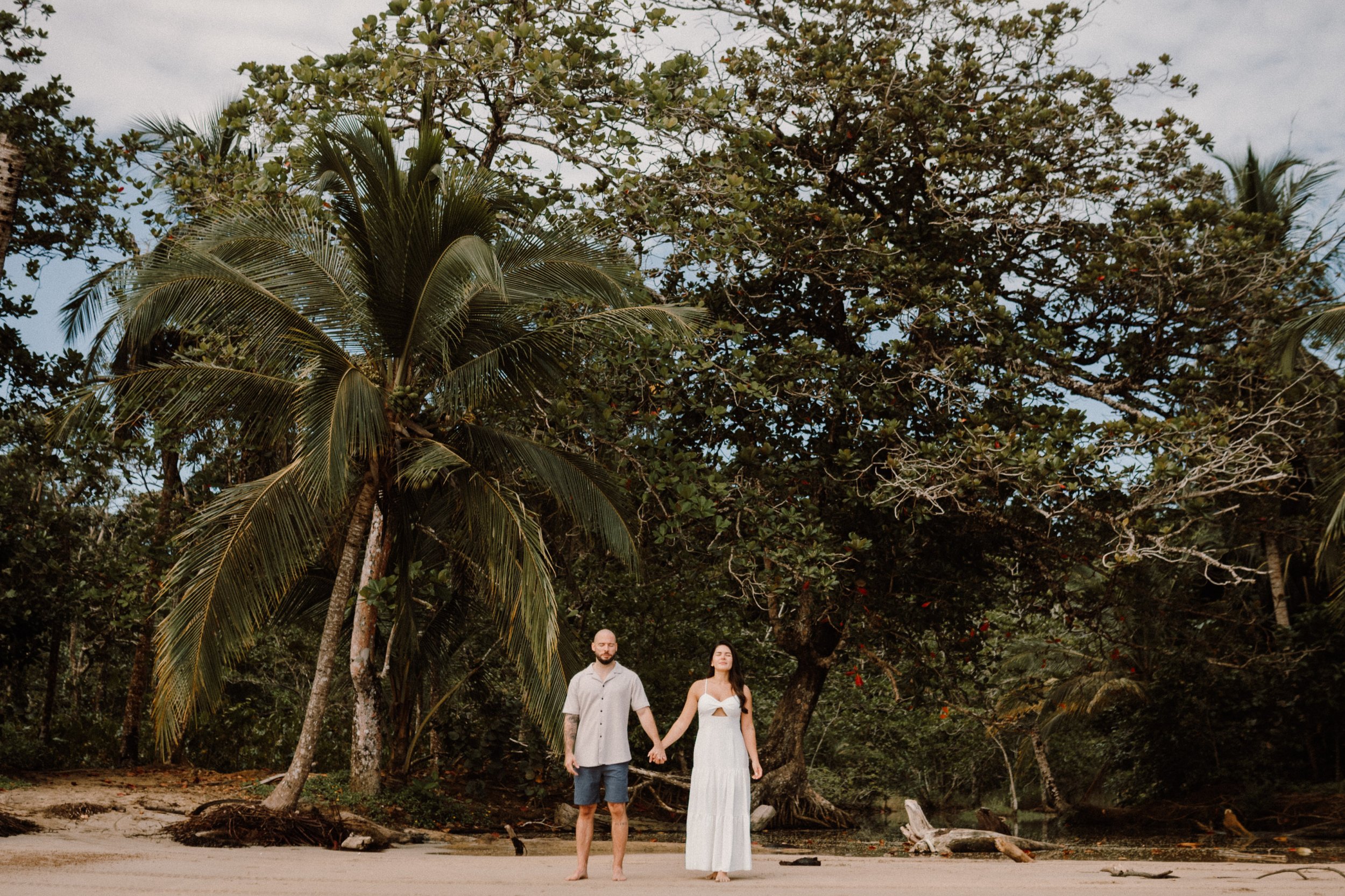 Una pareja sosteniéndose de la mano en una playa con palmeras y árboles tropicales.