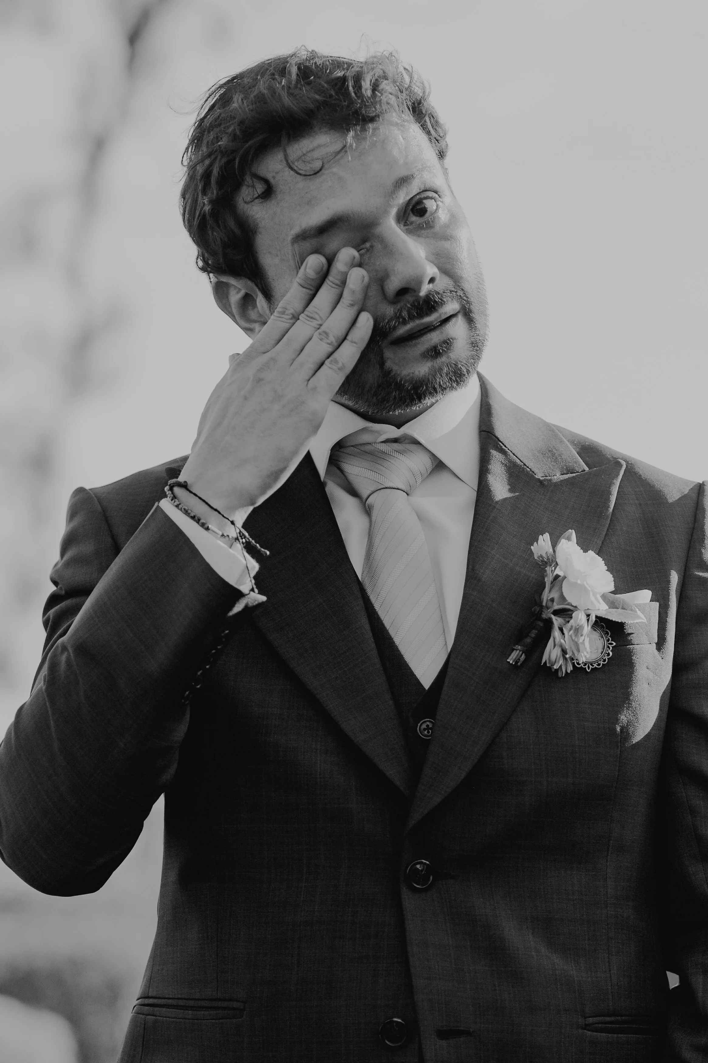 Hombre en traje con ramo en el saco, abrazando su rostro, con expresión de tristeza o emoción, en una escena en blanco y negro.