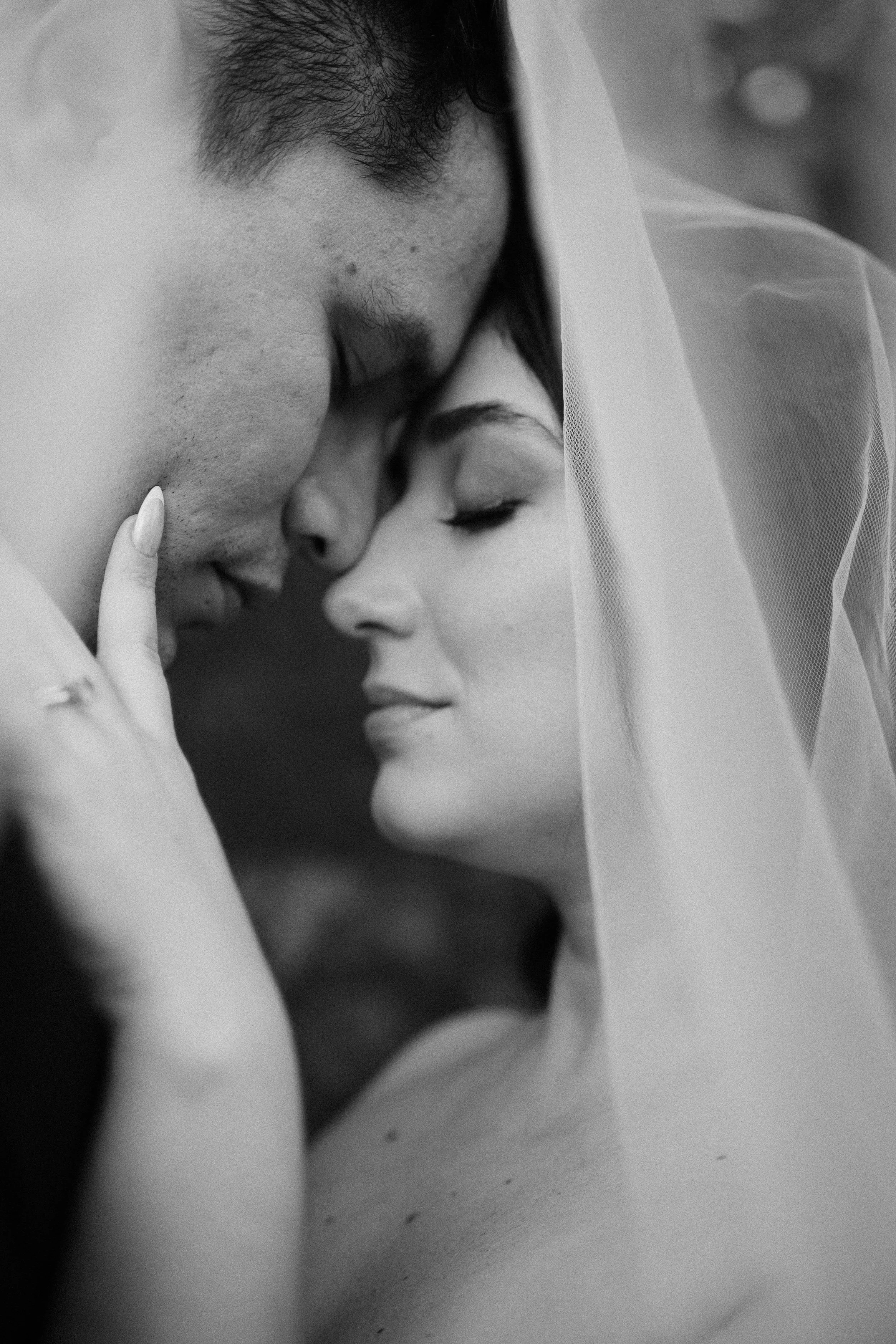 Una pareja en una escena romántica, con la pareja con los ojos cerrados, tocándose la cara y muy cerca, con un velo de novia en la mujer.
